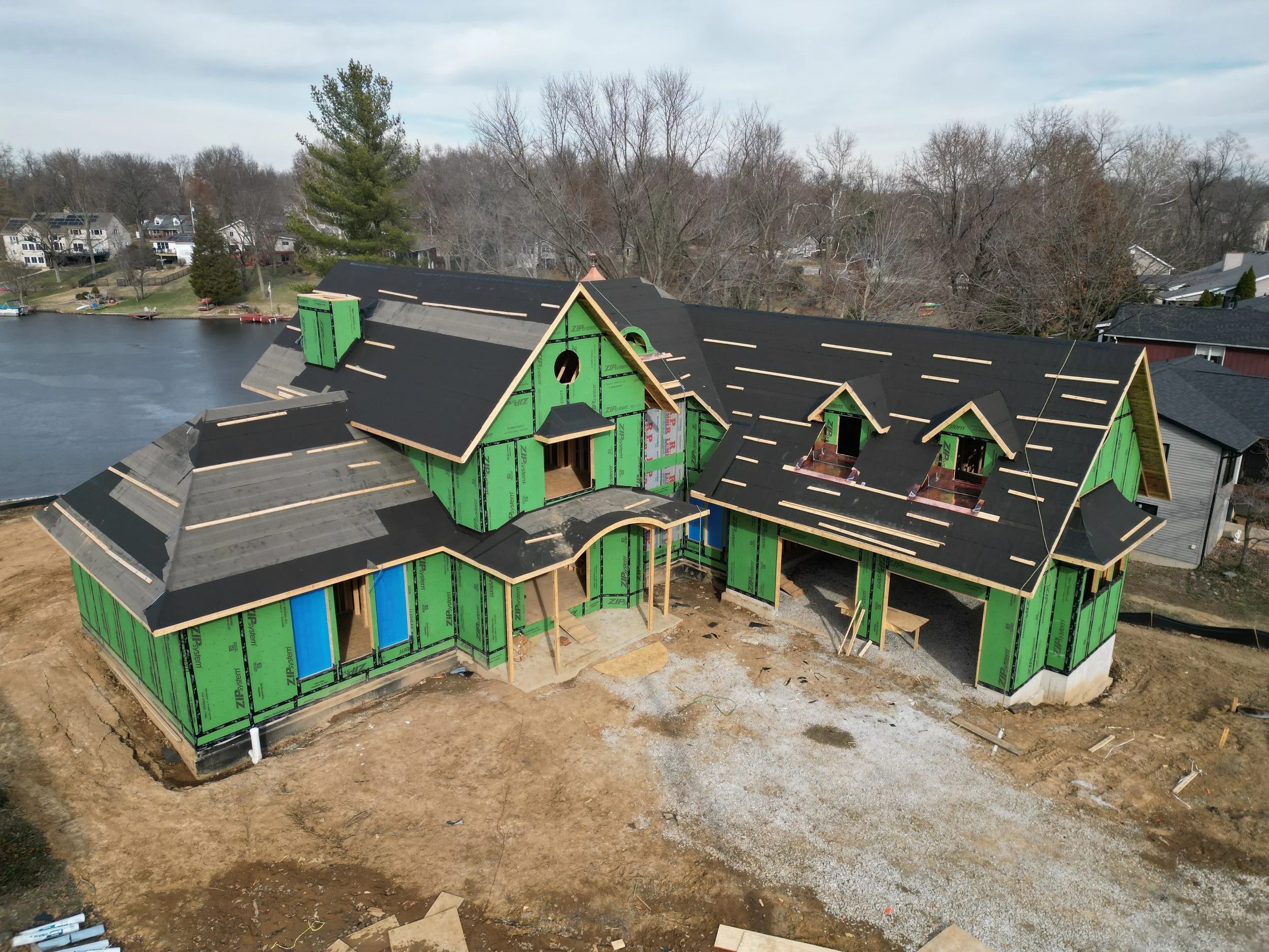 A house under construction, with green sheathing and black roofing, situated near a lake with a neighborhood in the background.