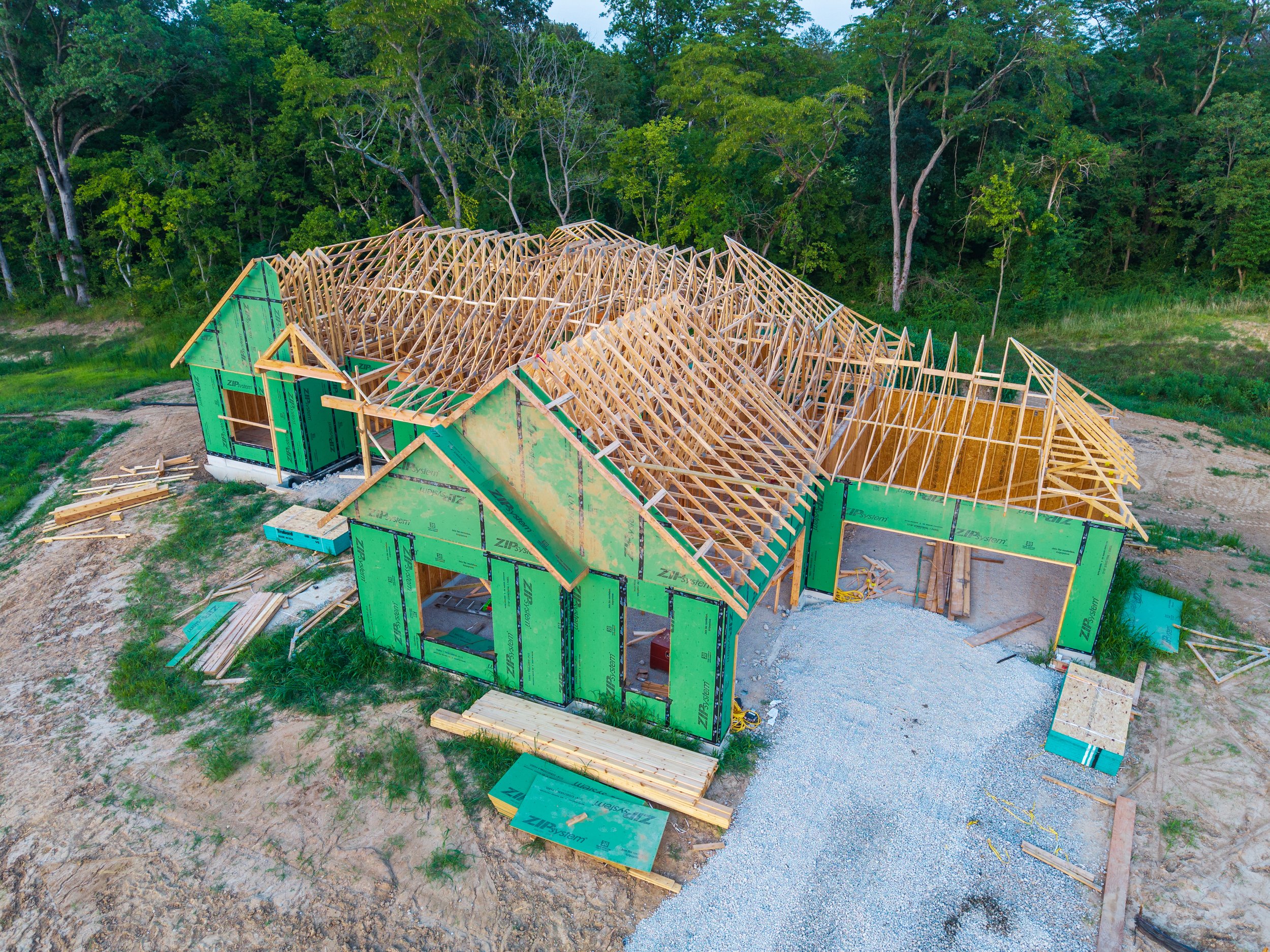 A house under construction with exposed wooden framing and green sheathing in a rural area with trees in the background.