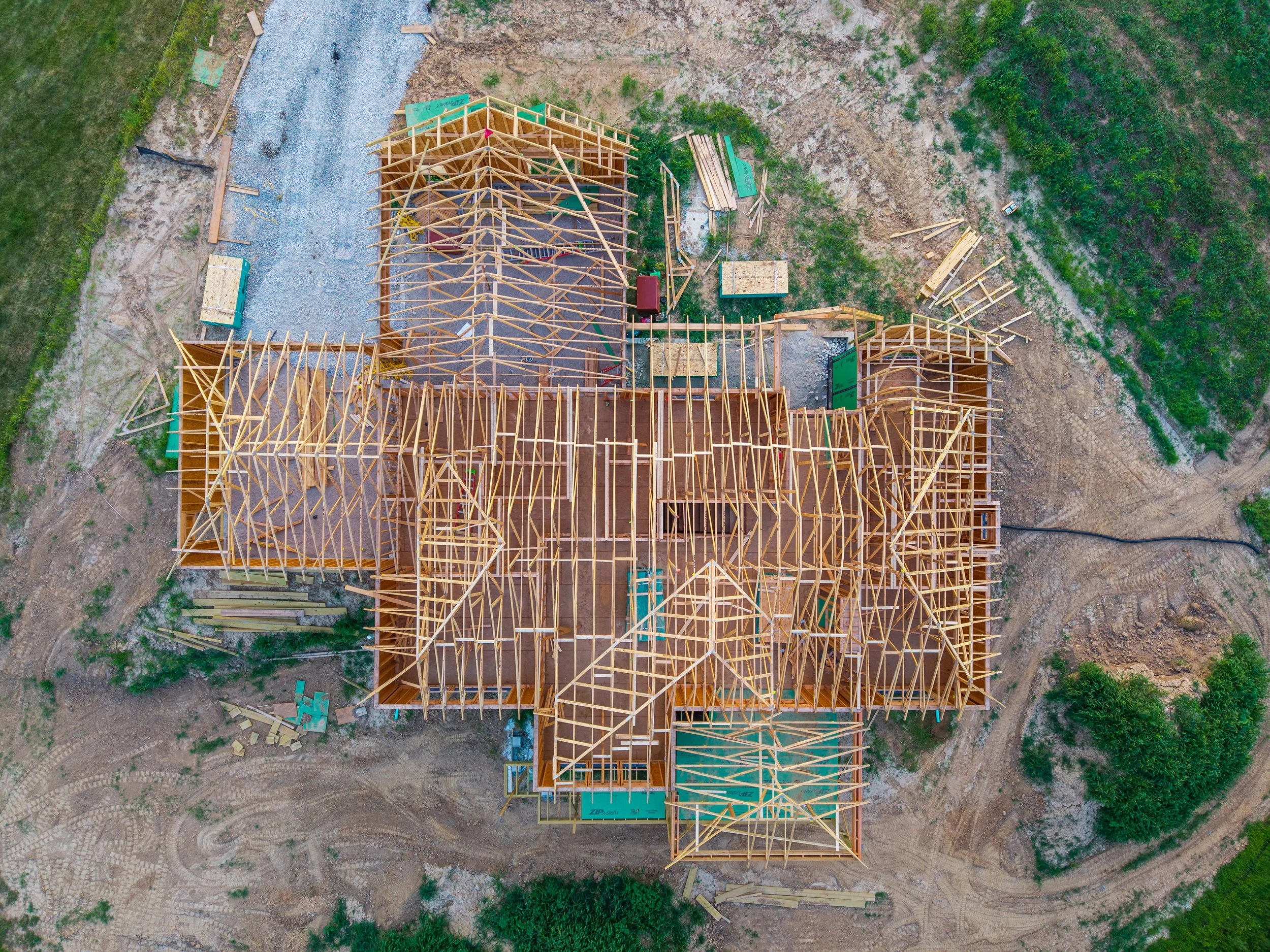 Aerial view of a house under construction with exposed wooden framework and green construction materials, surrounded by dirt and grass.