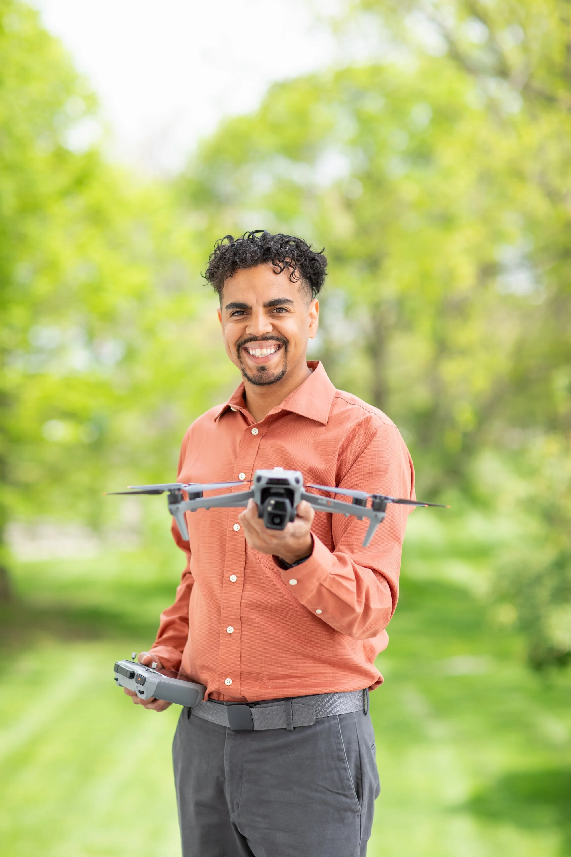 A man with curly hair smiling and holding a drone with a remote control, standing outdoors in a green park.