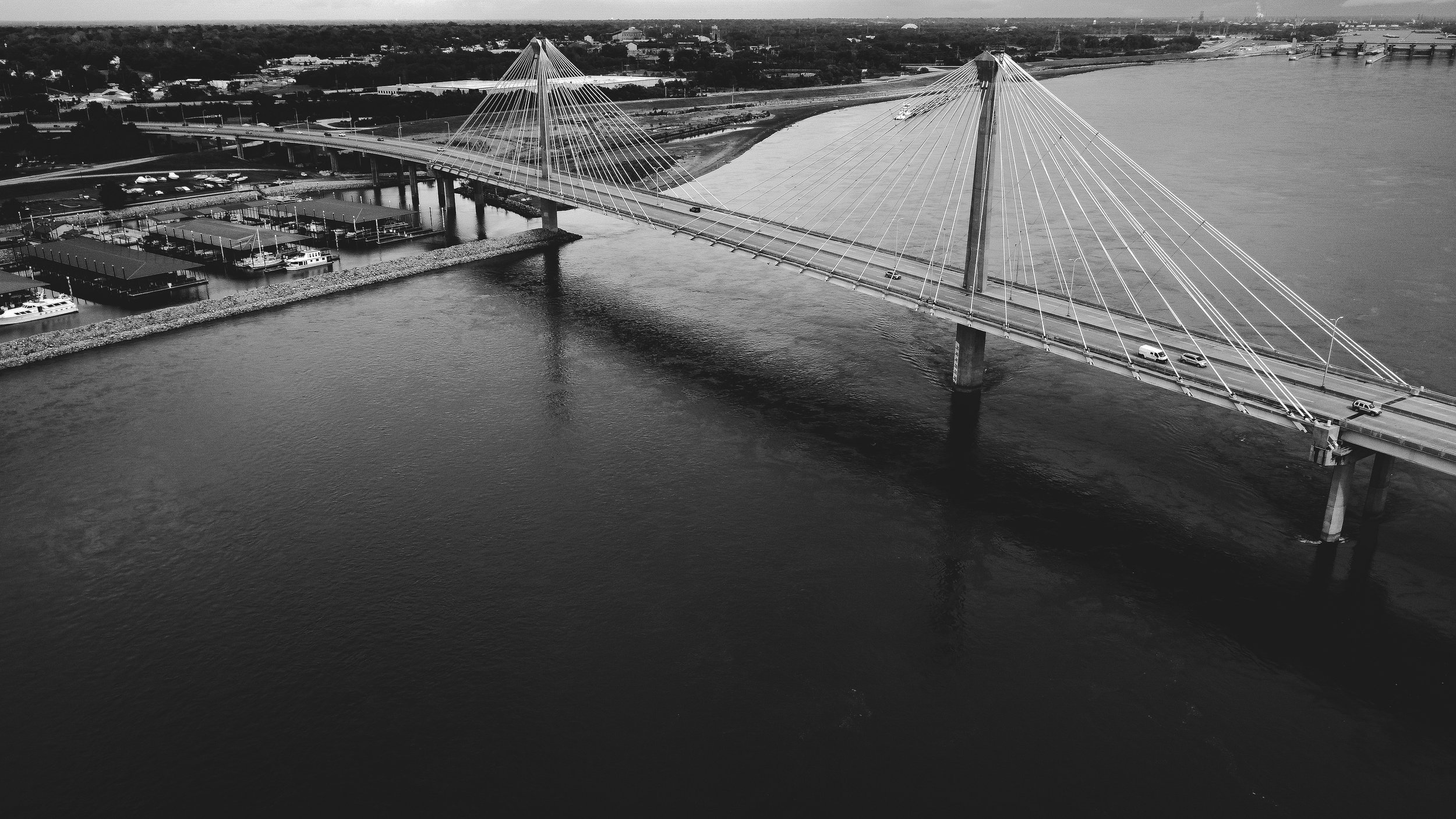 Aerial view of a cable-stayed bridge crossing a river, with cars traveling on it and boats docked at marinas nearby, in a cityscape