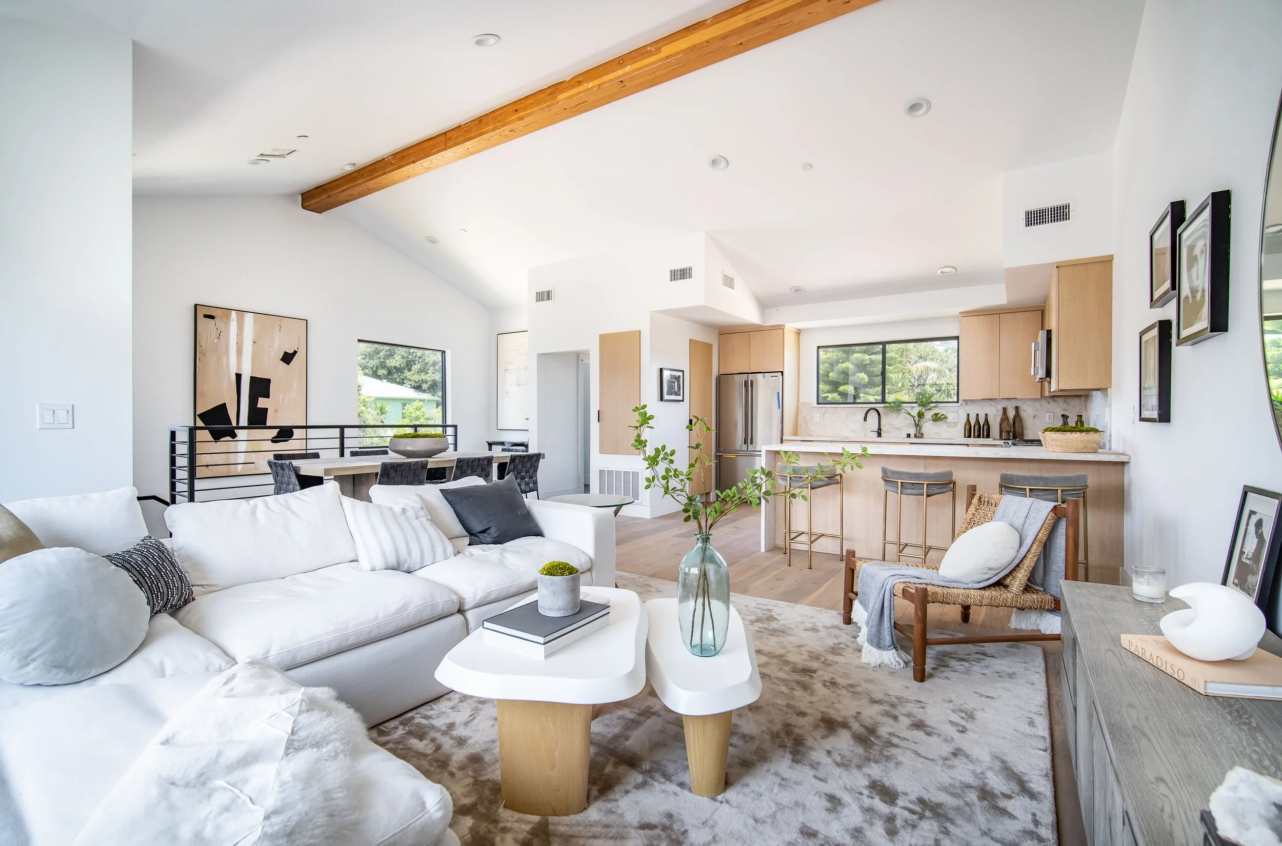Los Angeles staging and photography. Open-concept living room and kitchen with white walls, wooden ceiling beam, plush white sofa, armchair with blanket, modern coffee table, minimalist decor, and large windows overlooking greenery.