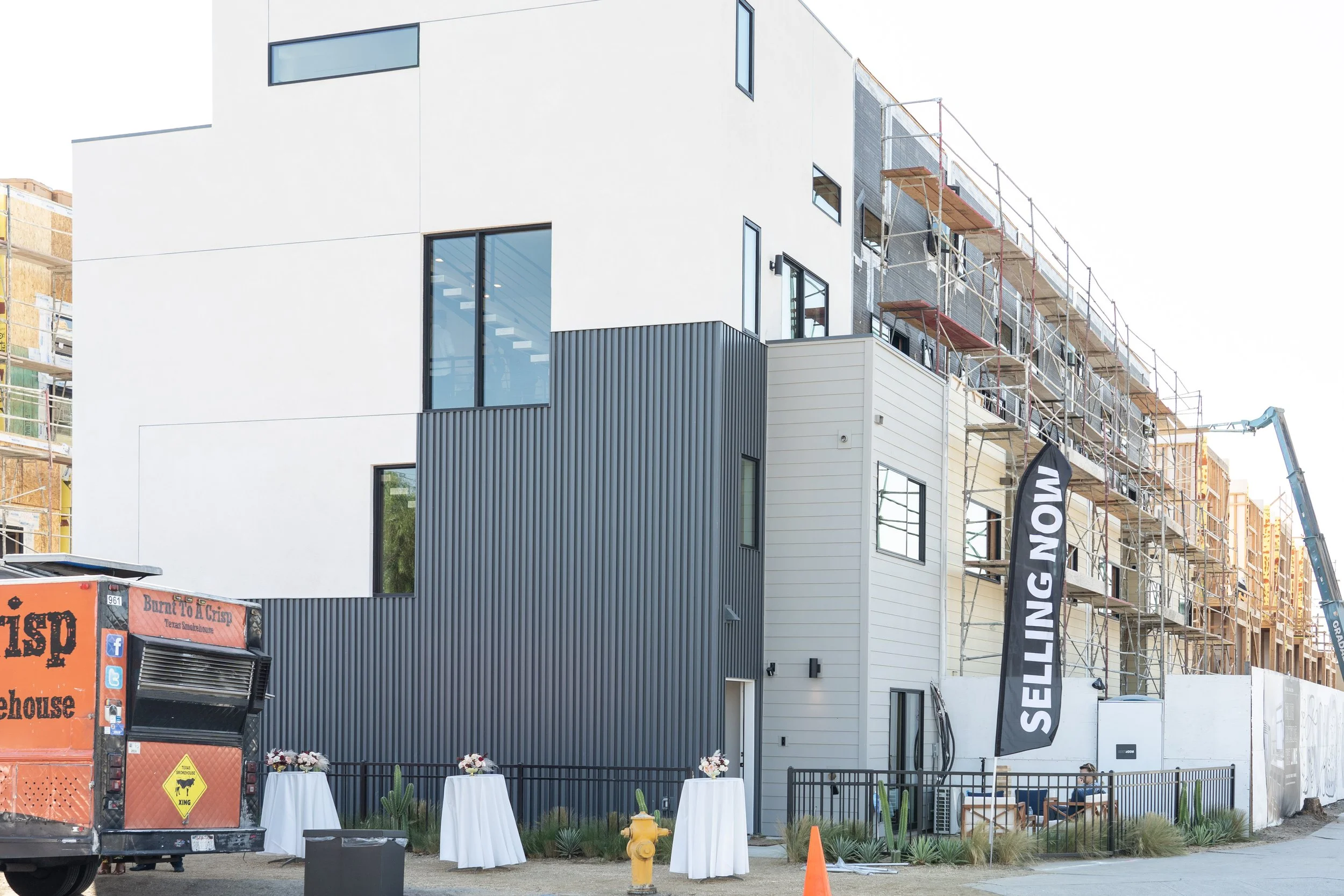 Construction site with a modern multi-story building in progress, scaffolding on the right, a black flag with white text 'SELLING NOW,' and a food truck on the left.