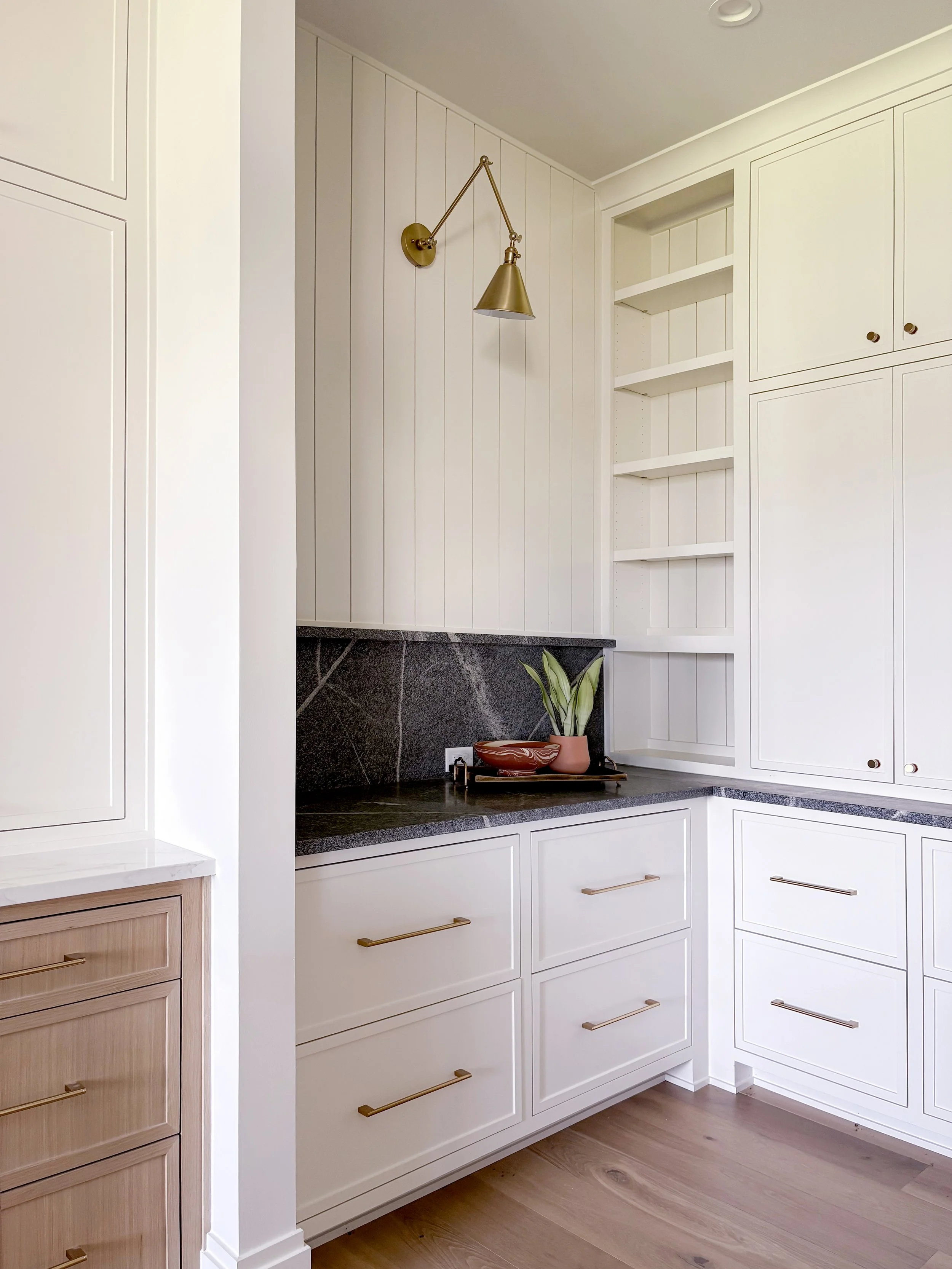 Kitchen and butler pantry corner with white cabinetry, black granite countertop, open shelves, and a brass wall-mounted lamp.