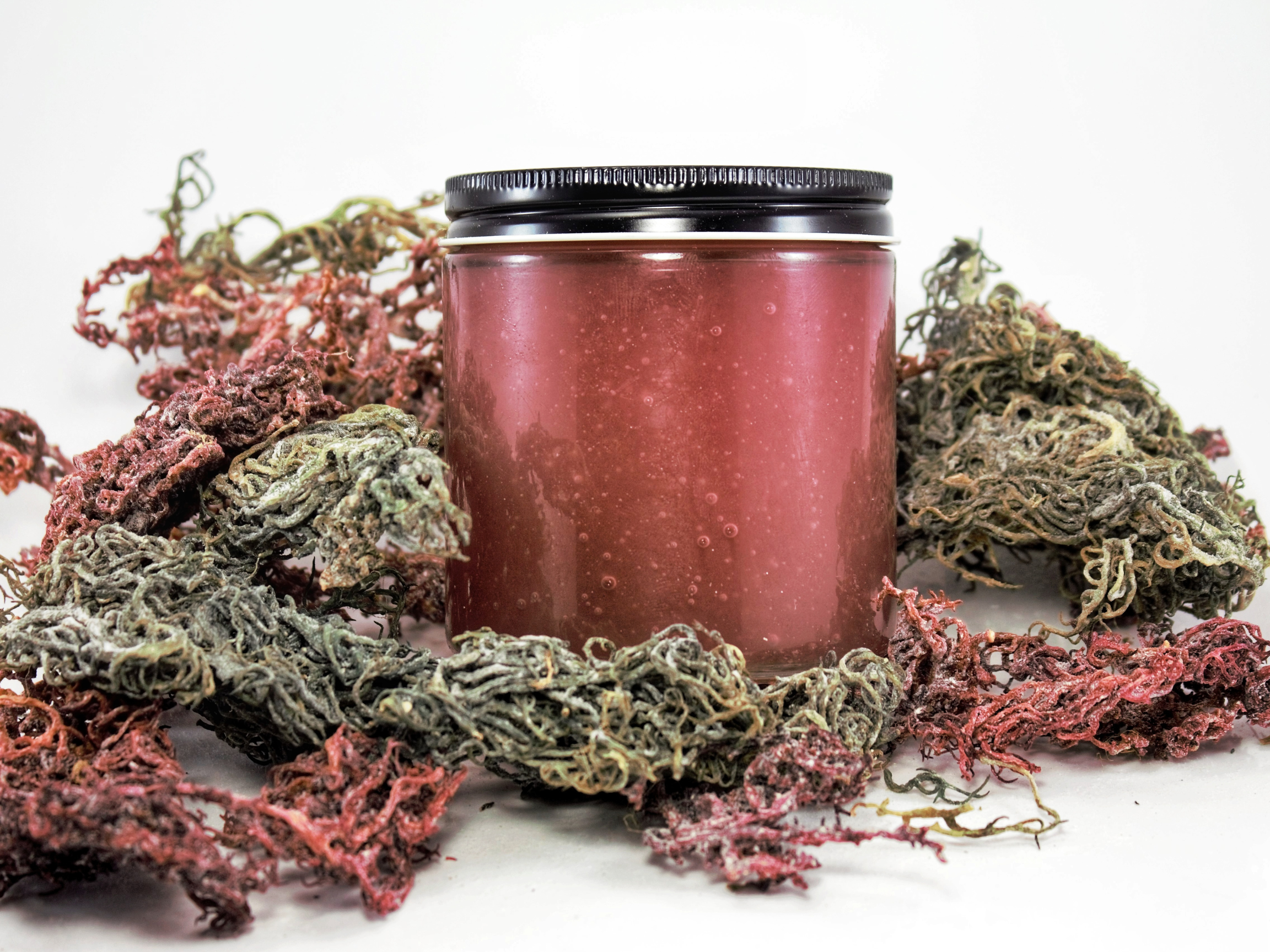 A pink jar with a black lid surrounded by dried purple, green, and pink marijuana buds on a pink background.