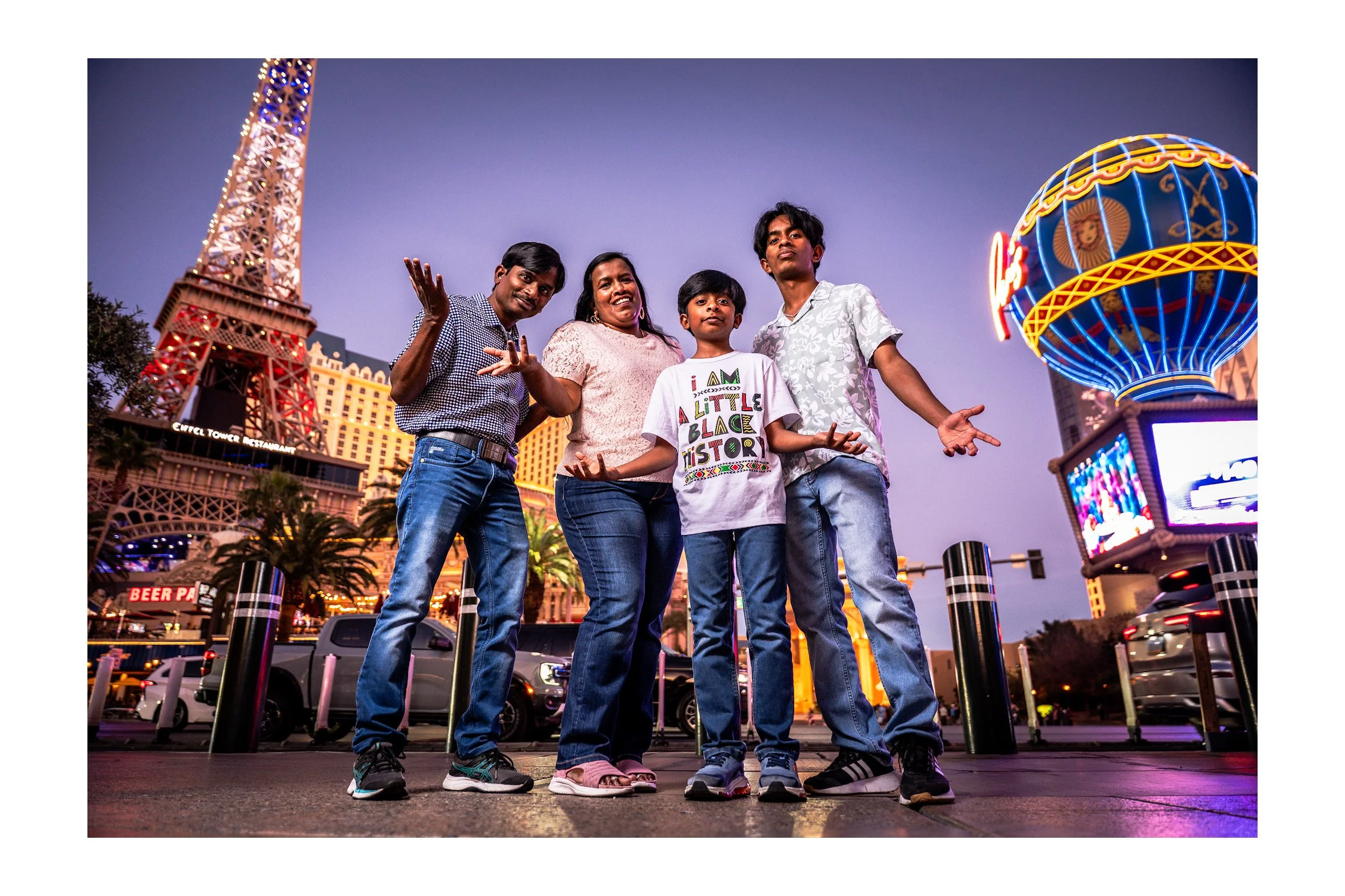 Four people, a man, woman, and two boys, standing in front of New York-New York Hotel & Casino with the replica Brooklyn Bridge and the Eiffel Tower in the background, at dusk in Las Vegas.