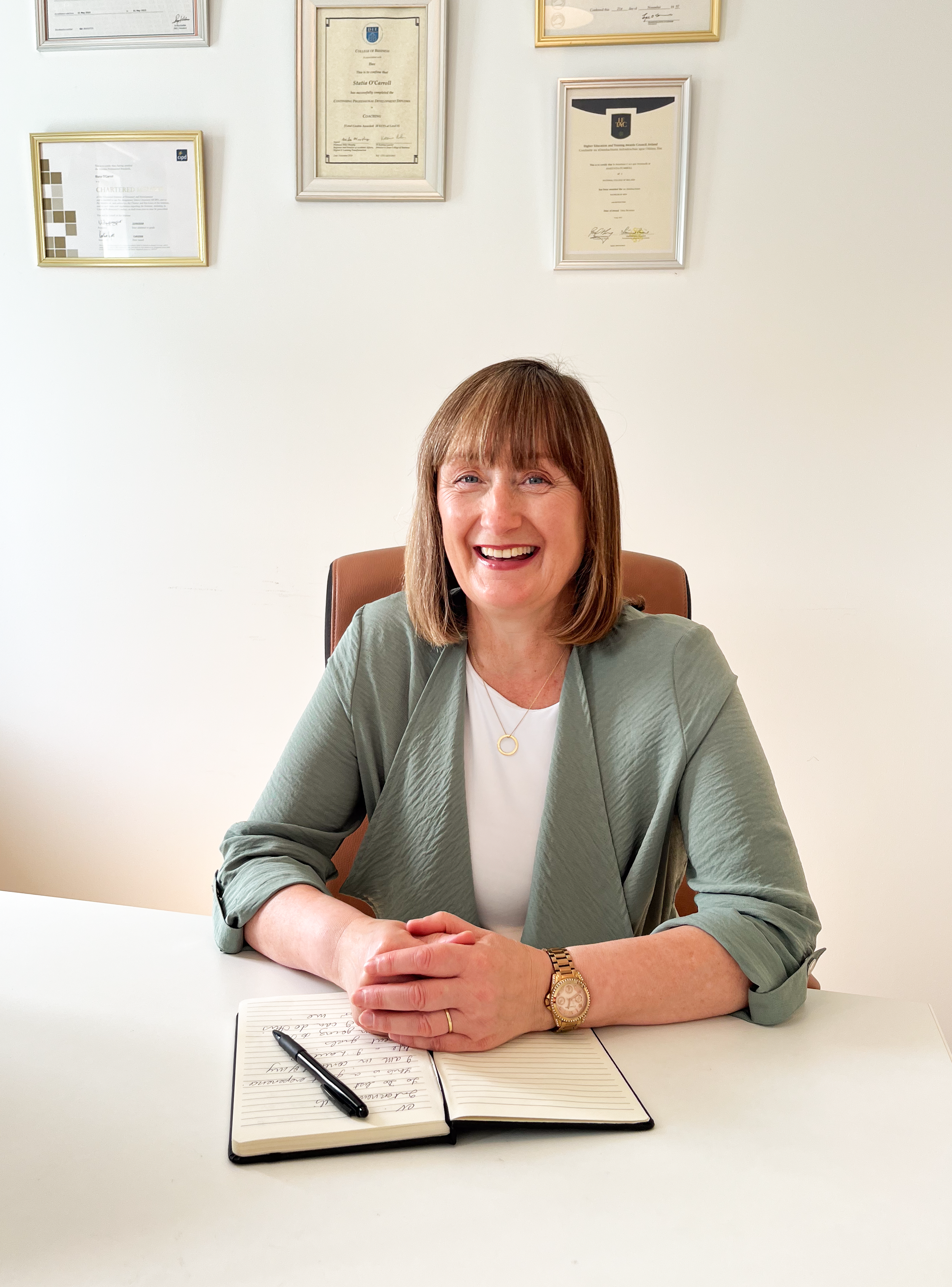 A woman with shoulder-length brown hair, wearing a light green blazer and a white top, sitting at a white desk with a black pen and an open notebook. She is smiling and sitting in front of a white wall decorated with framed certificates or diplomas.