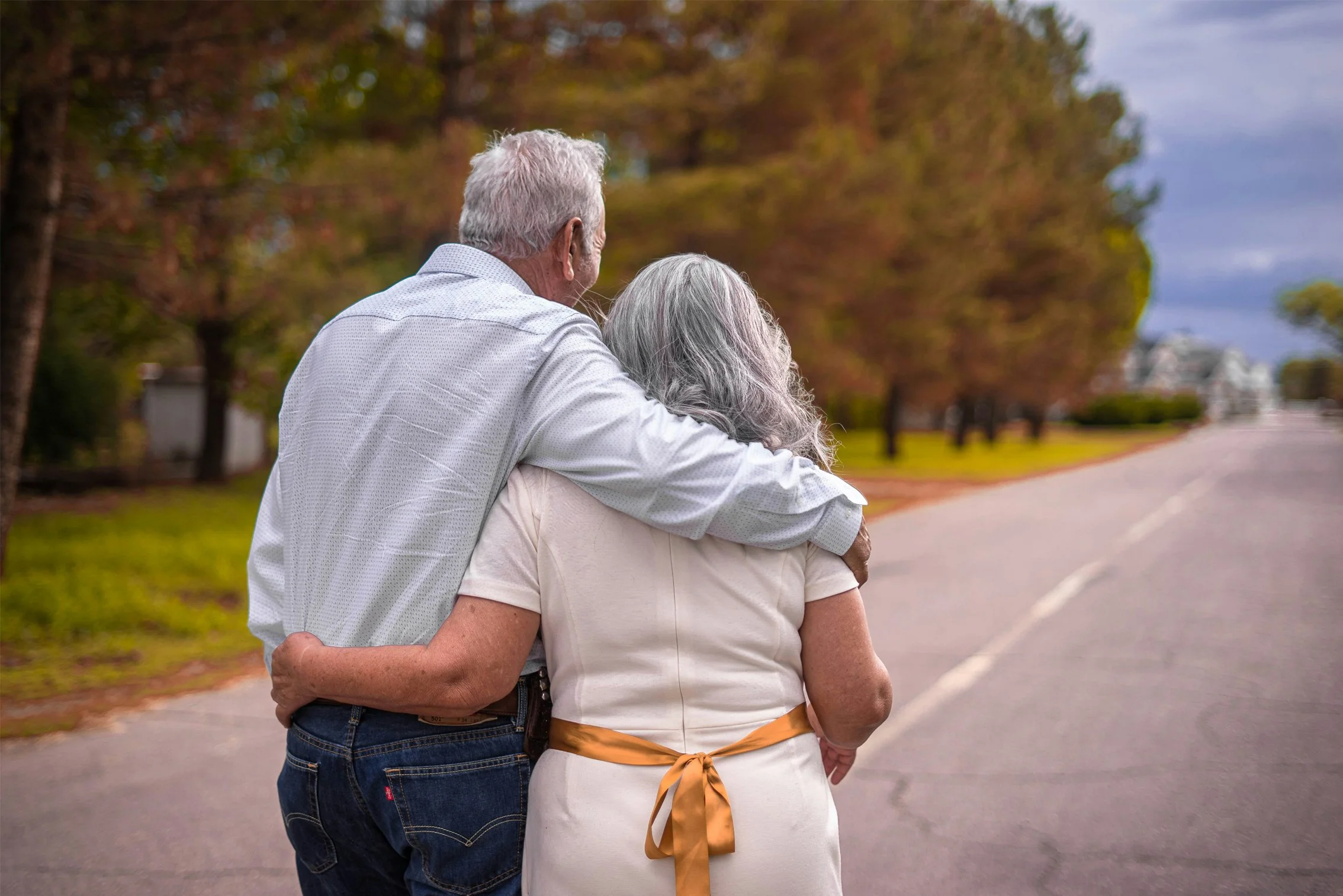 older man with grey hair has arm around older woman with grey hair walking down the road.
