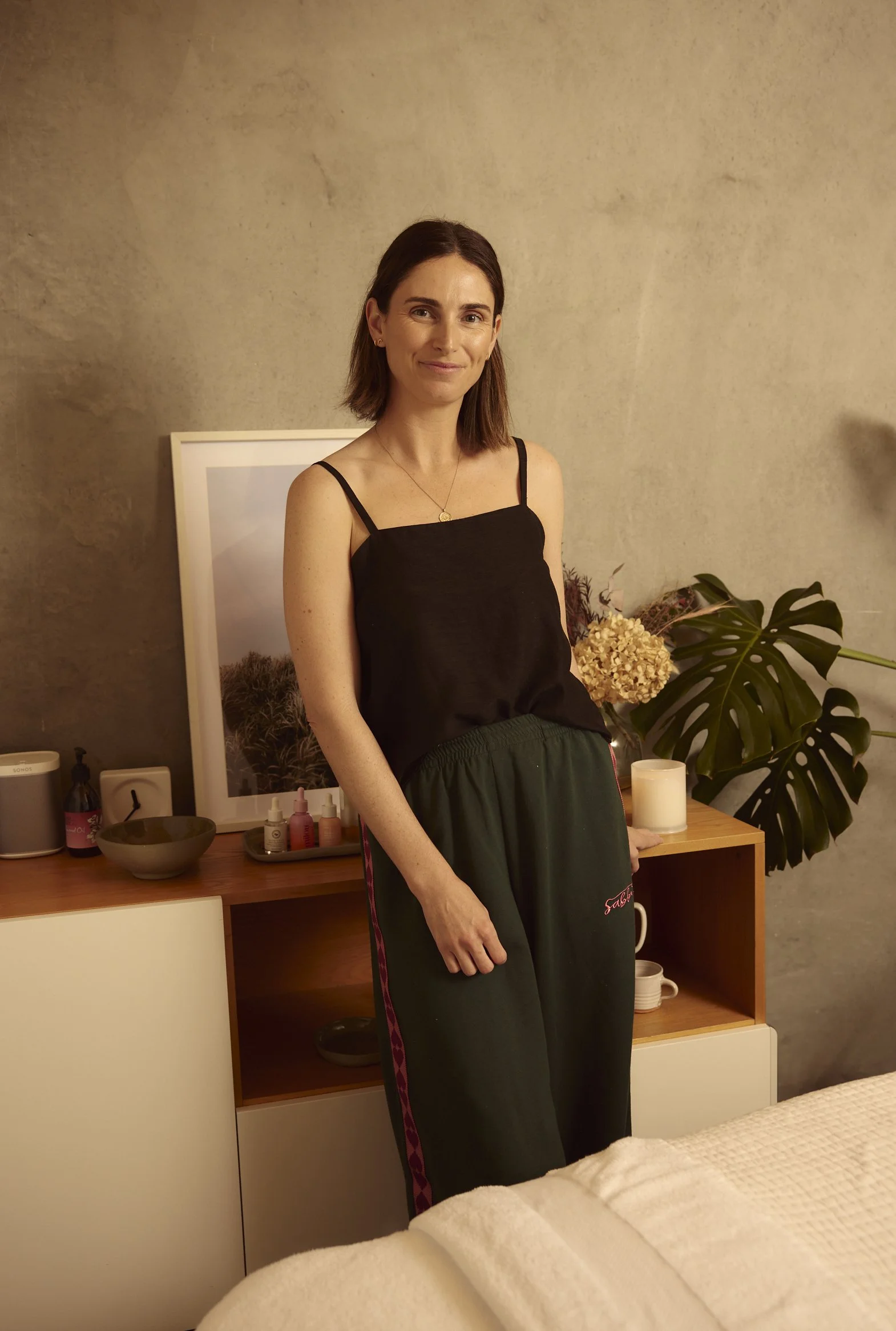 A woman with shoulder-length brown hair wearing a black camisole top and dark green pants standing in a room with a textured beige wall, a wooden cabinet with various items including bottles, a large leafed plant, a round candle, and a framed picture.