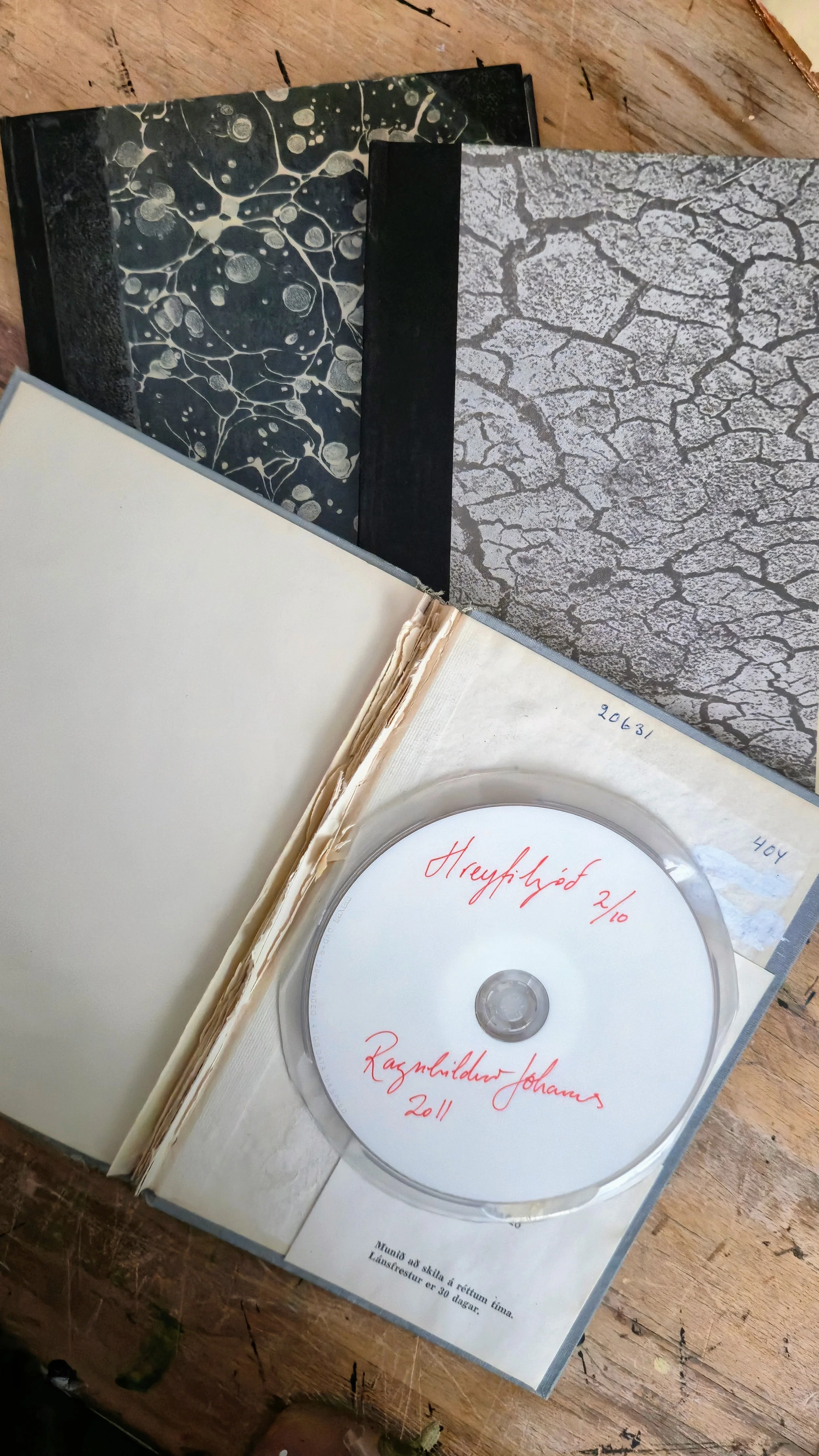 An open photo album with handwritten text on the right page, alongside a CD with red handwritten text. The background includes patterned papers or covers, one black with white abstract patterns and another gray with a crackle pattern, resting on a wooden surface.