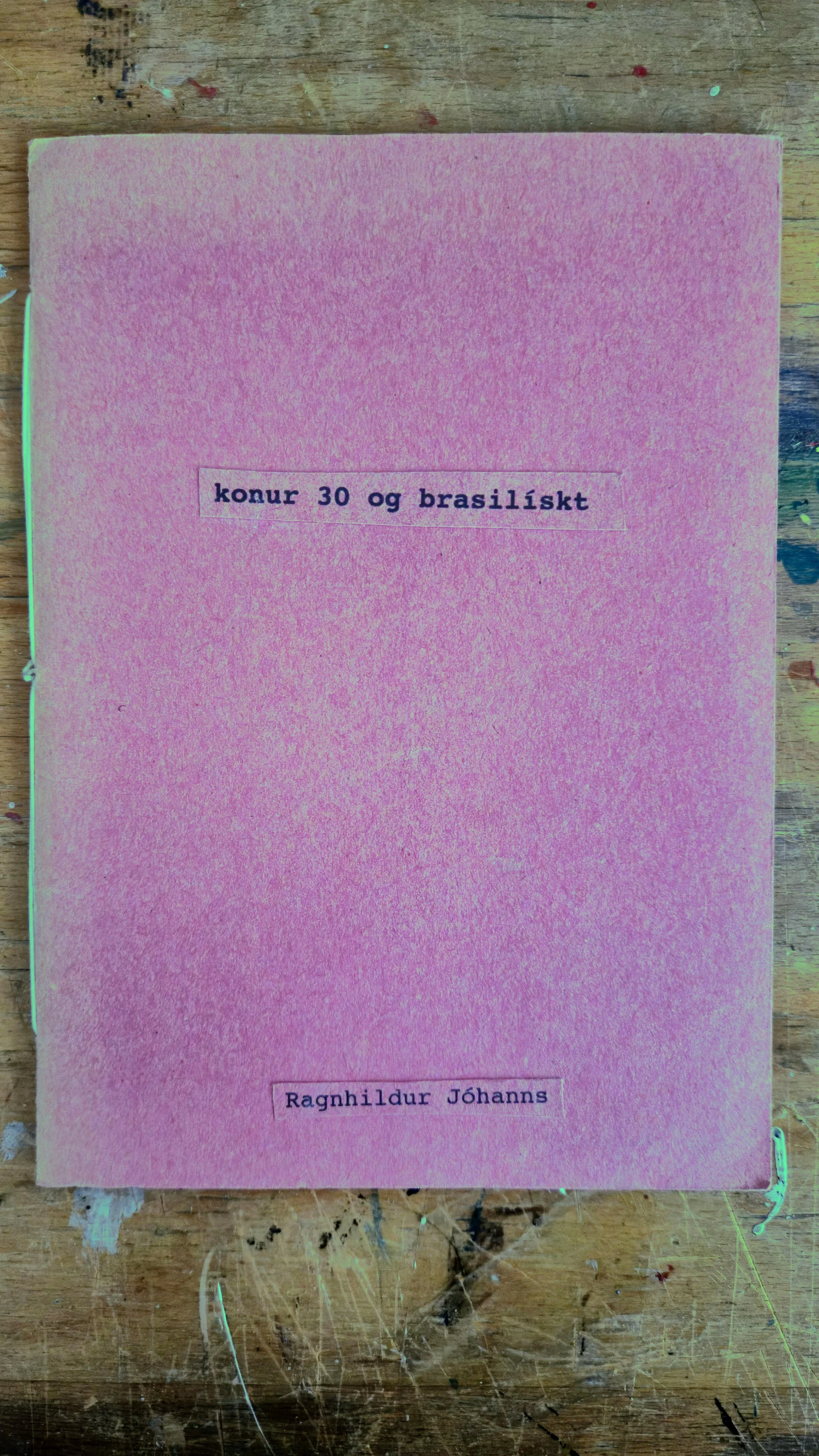 Pink book cover with typed text in Icelandic that reads 'konur 30 og brasilískt' and the author's name 'Ragnhildur Jóhanns' printed on it, placed on a worn wooden surface.