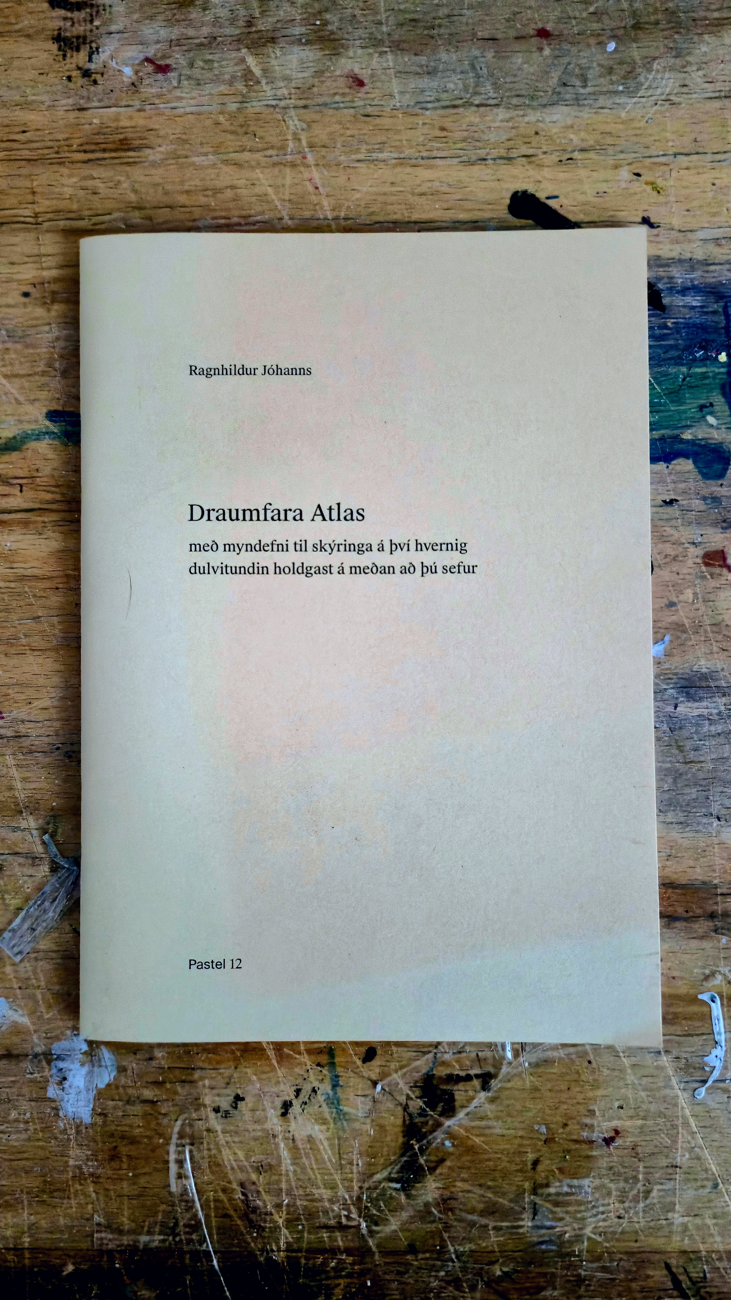 A book titled 'Draumfara Atlas' by Ragnhildur Jóhanns resting on a wooden surface with paint splatters.