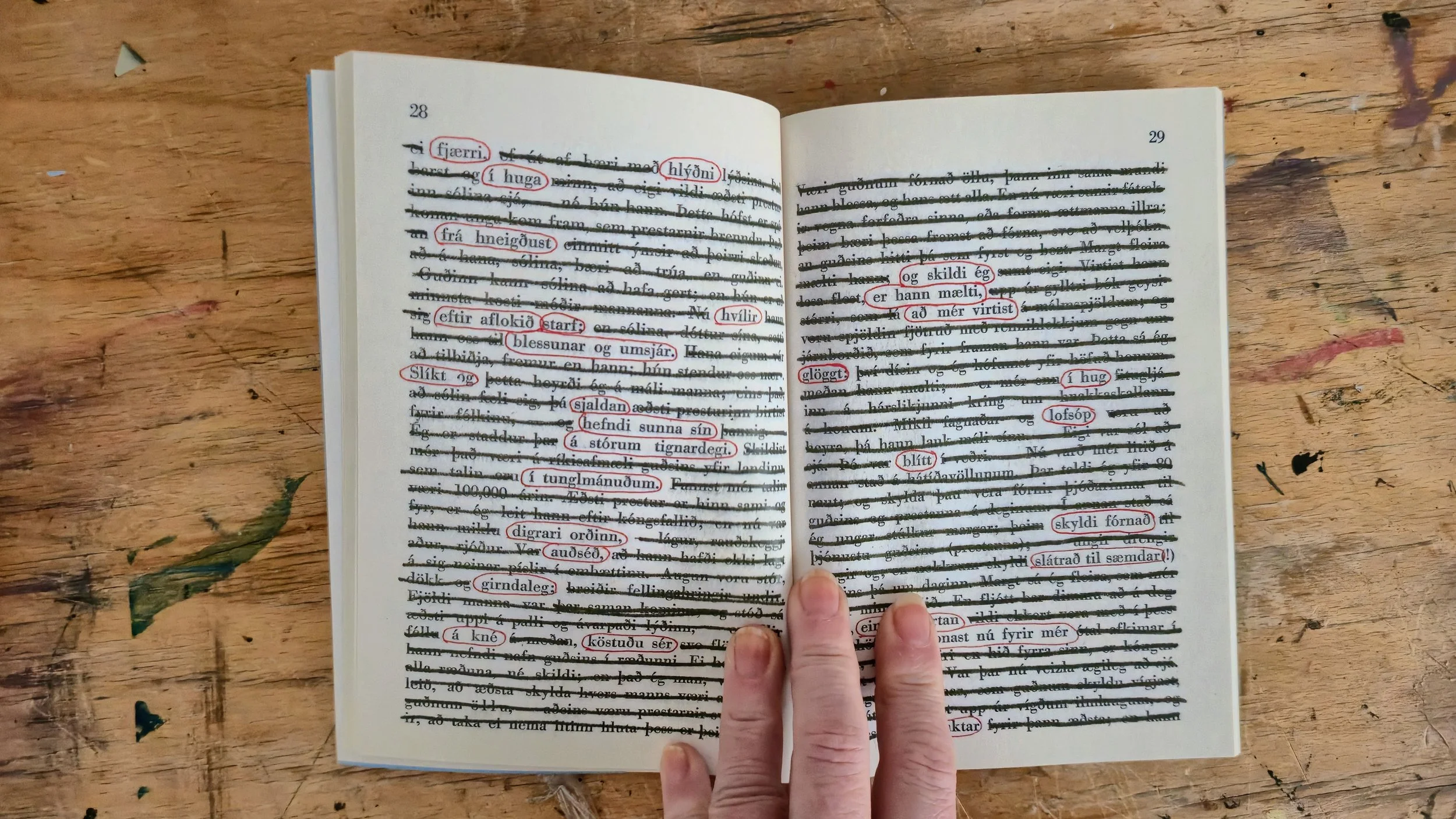 Open book with blacked-out text and Icelandic words circled in red, held by person's fingers, on a wooden surface.