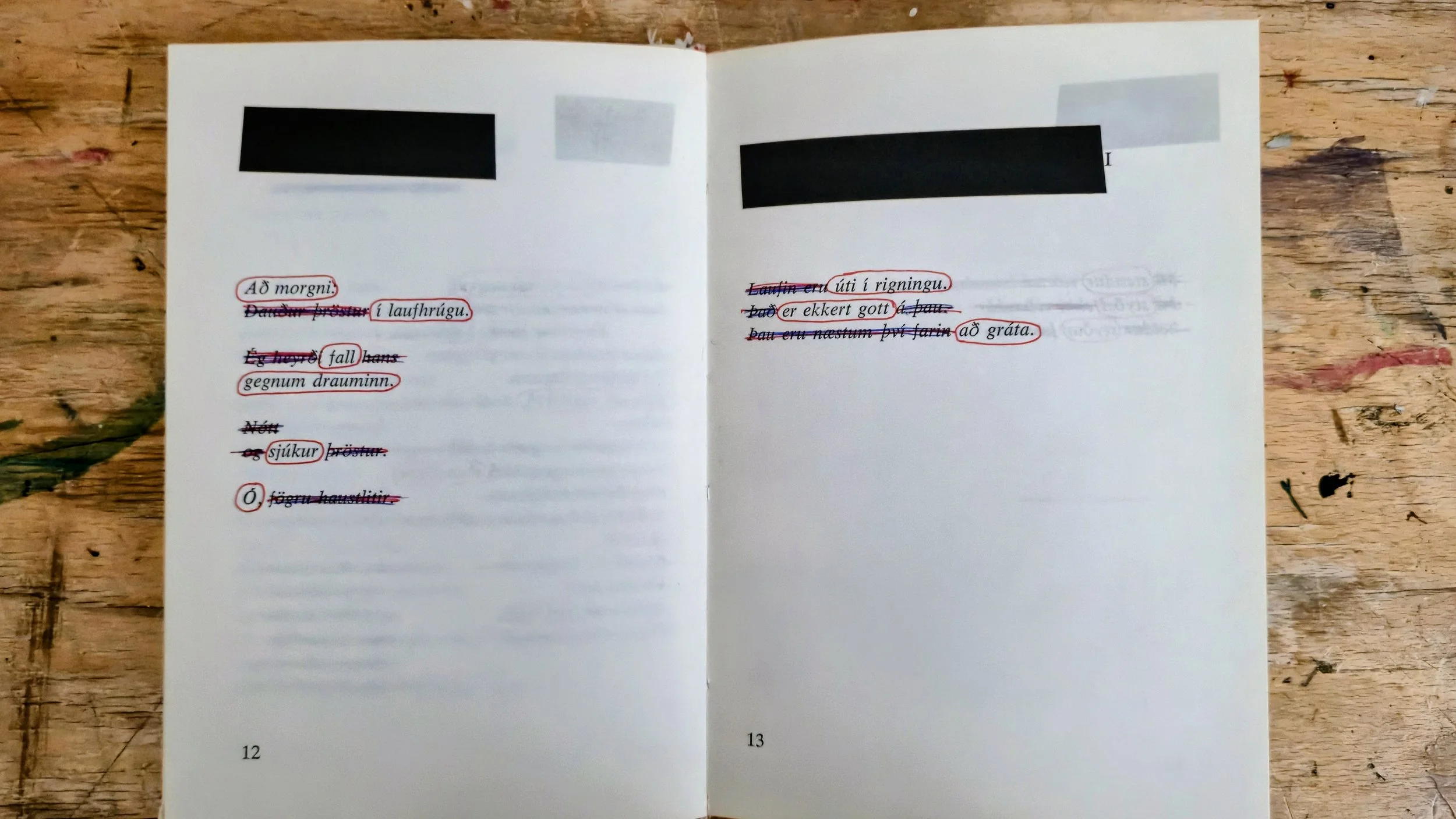 Open book with handwritten notes in Icelandic, pages 12 and 13, on a wooden surface. The notes are underlined and crossed out with red, blue, and purple ink.
