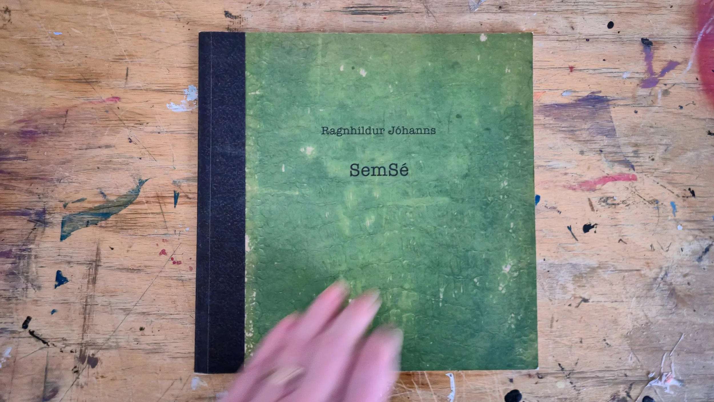 A green book titled 'Ragnhildur Jóhanns Semsé' lying on a wooden table with colorful paint splatters; a person's fingers are partially visible at the bottom.