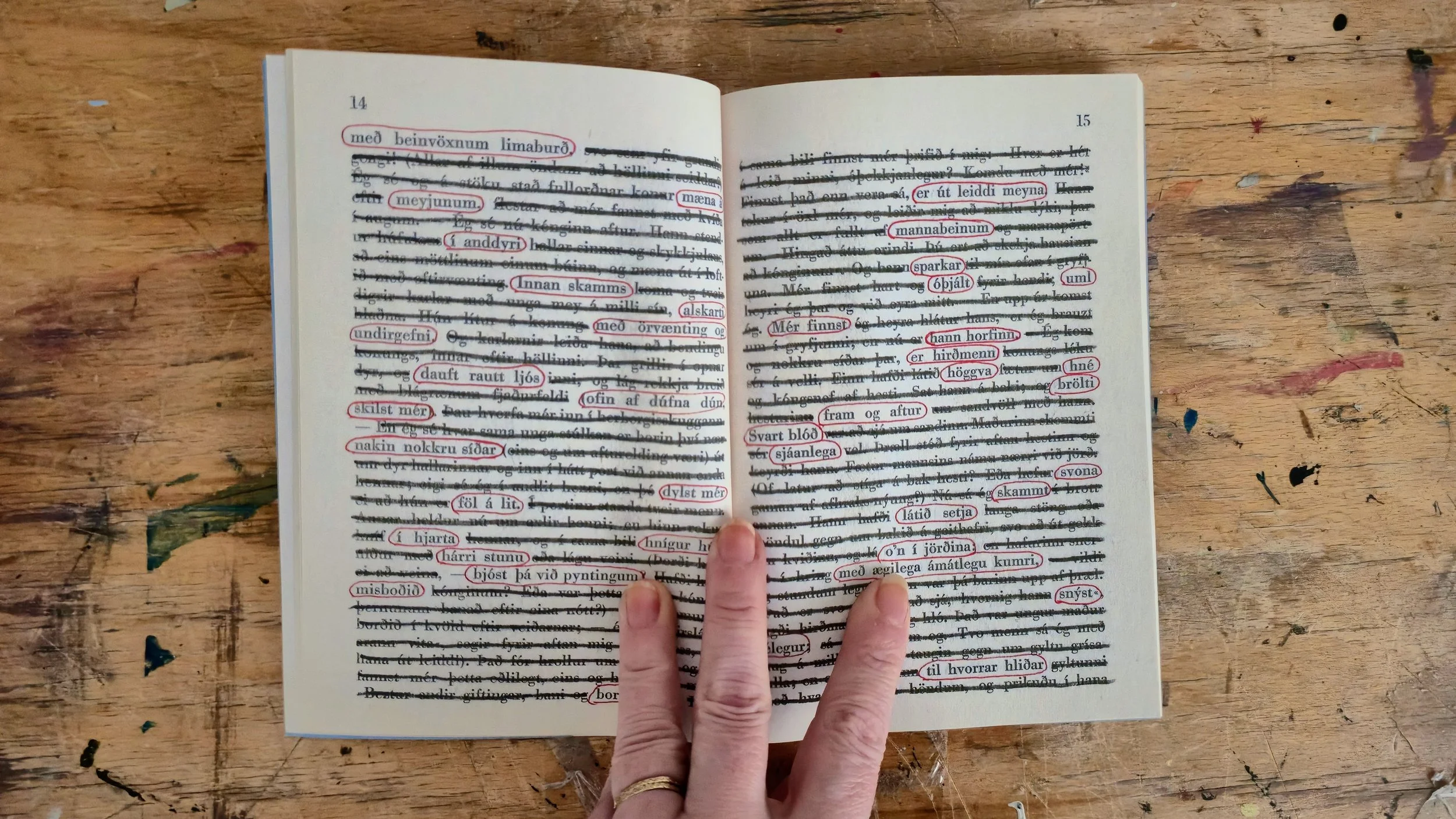 Open book with Icelandic text and red markings, held by a person's hand, on a wooden surface with marks and paint splatters.