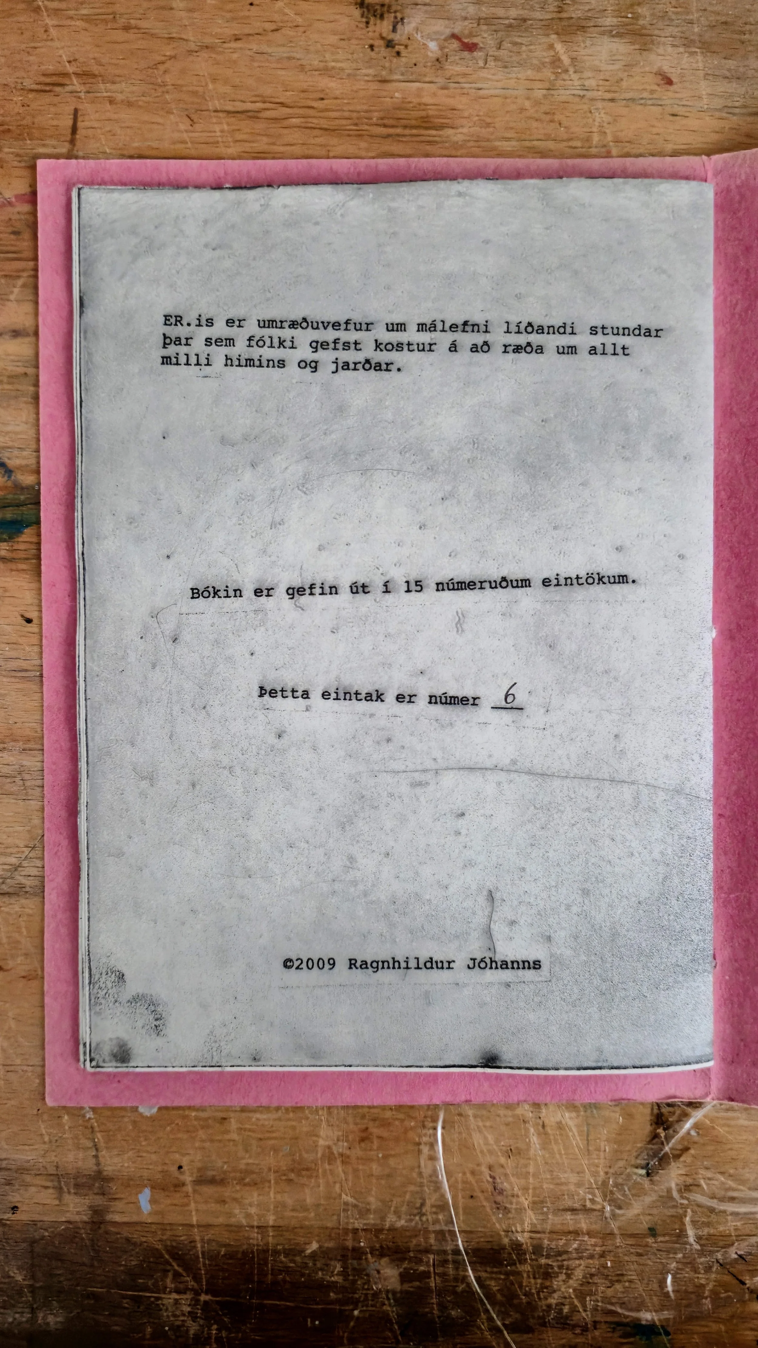 A black-and-white printed page with Icelandic text, placed on a pink background on a wooden surface.