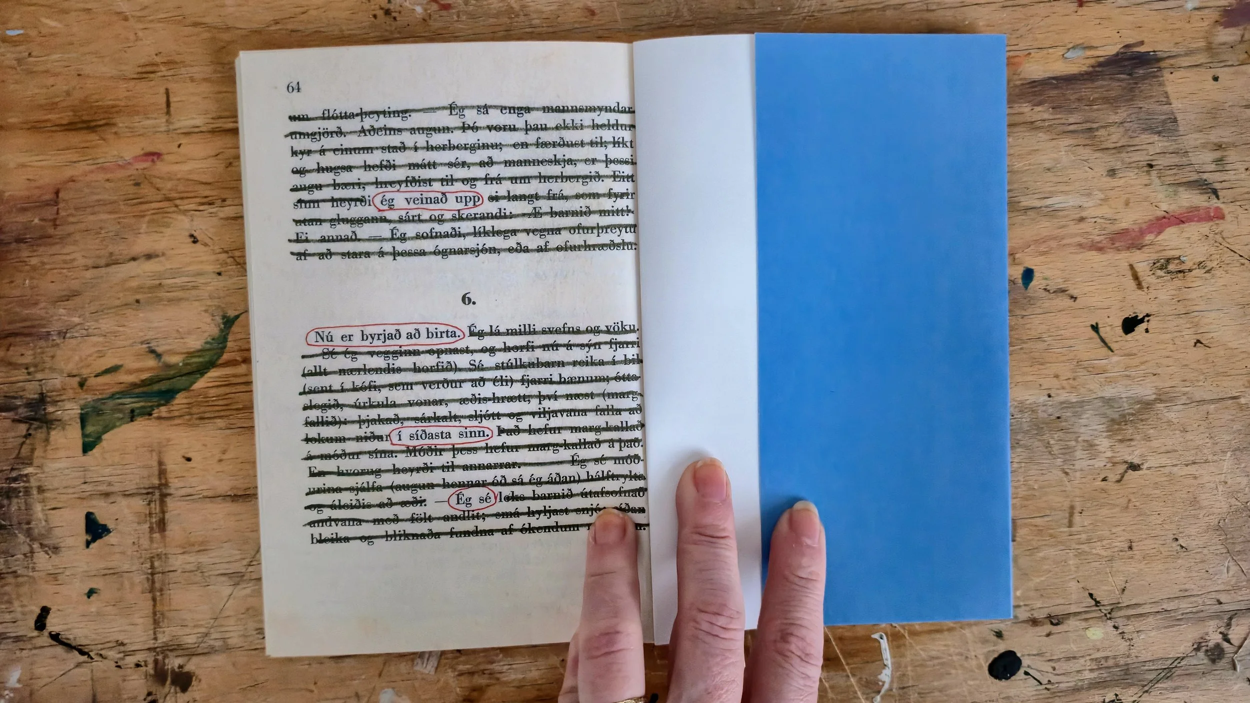 Open book resting on a wooden table, with a person's hand holding the bottom right corner. The book page shows Icelandic text with some phrases circled in red ink.
