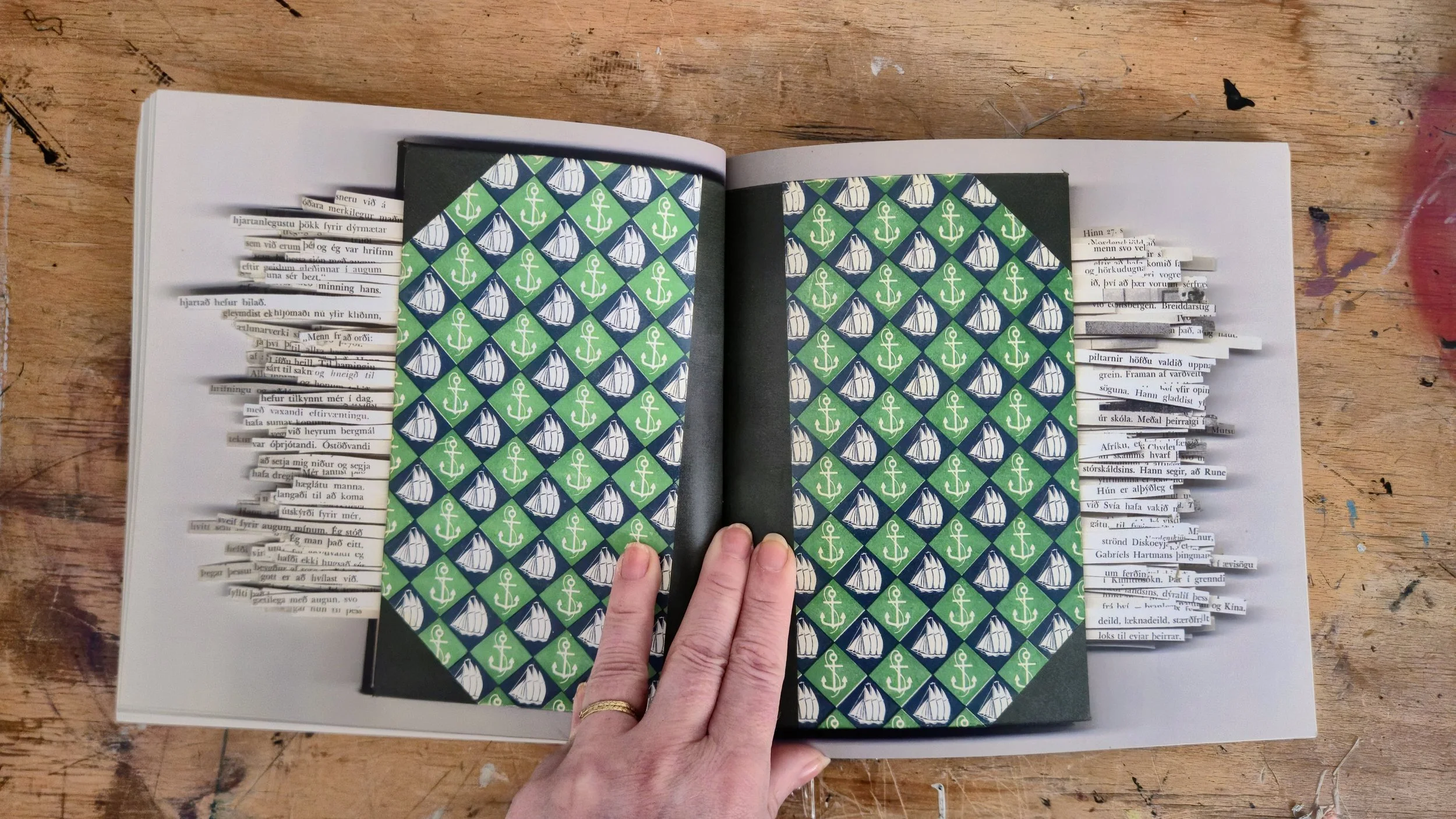 Open book on a wooden table, with pages containing dived newspaper strips on the sides and patterned paper with sailboats and anchors in the center of the pages.