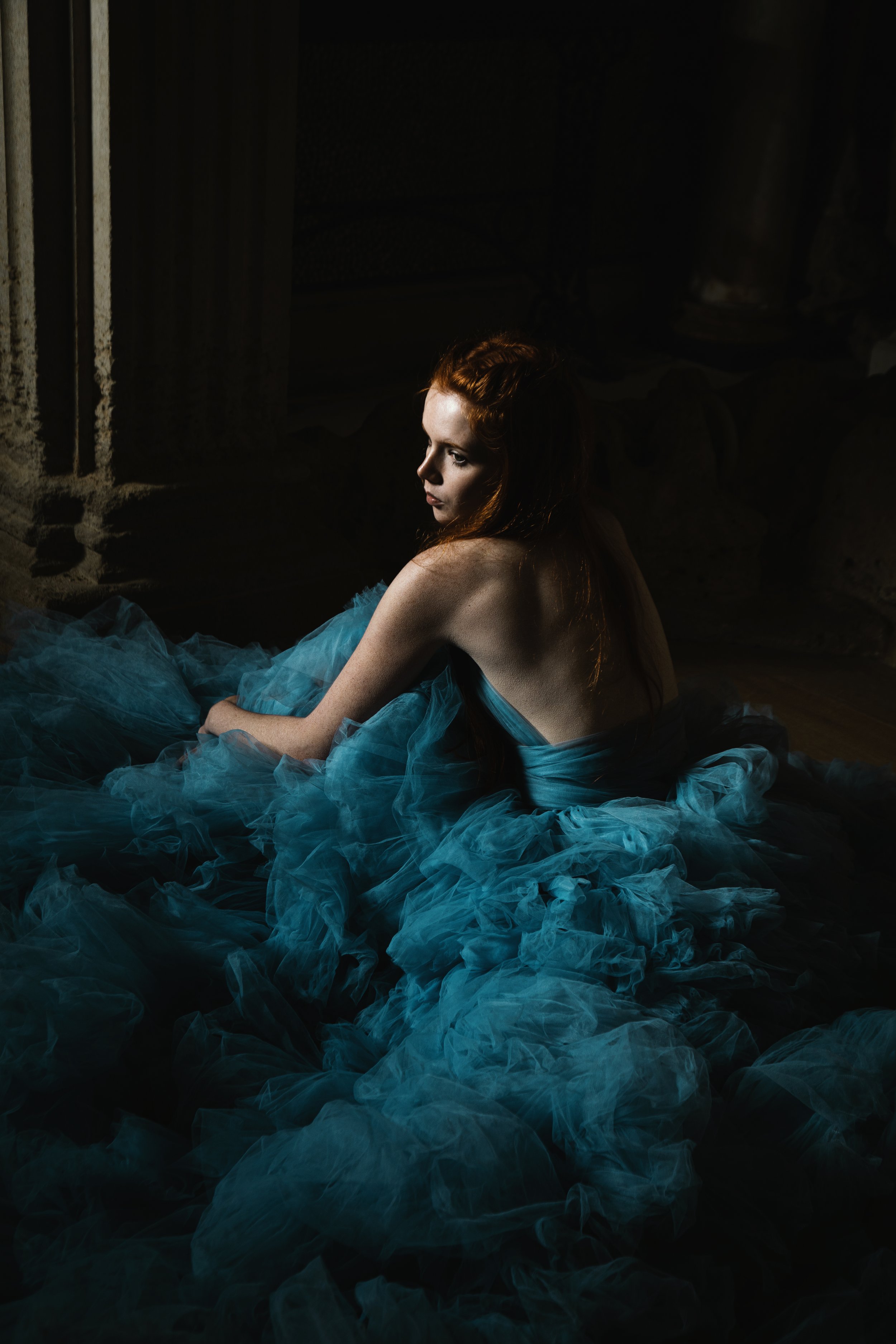 A woman with red hair sitting in a dark room, wearing a blue tulle dress, with soft lighting illuminating her face and upper body.
