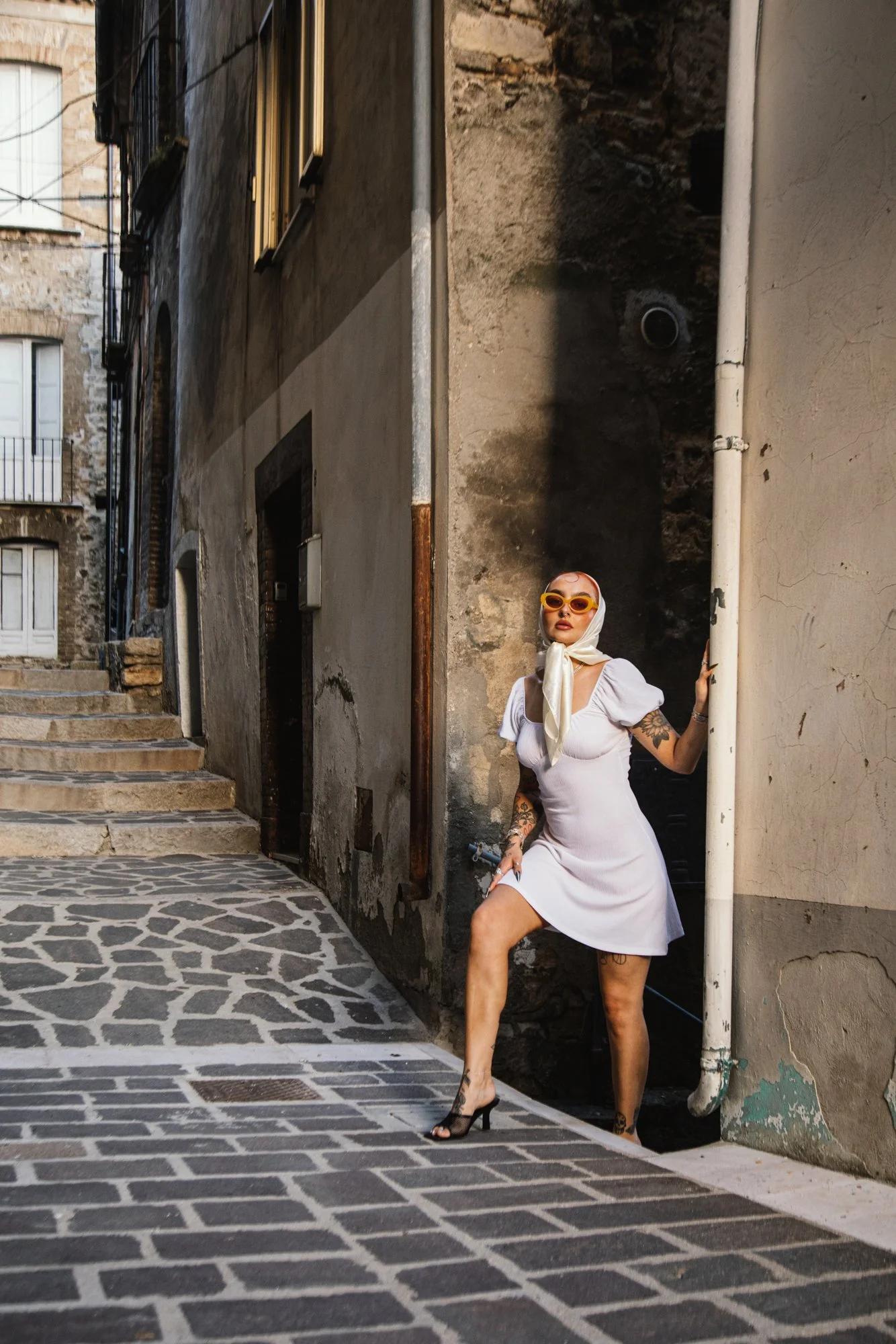 A woman in a white dress with tattoos, wearing high heels and yellow sunglasses, standing against a corner wall in an alleyway with old buildings and a cobblestone sidewalk.