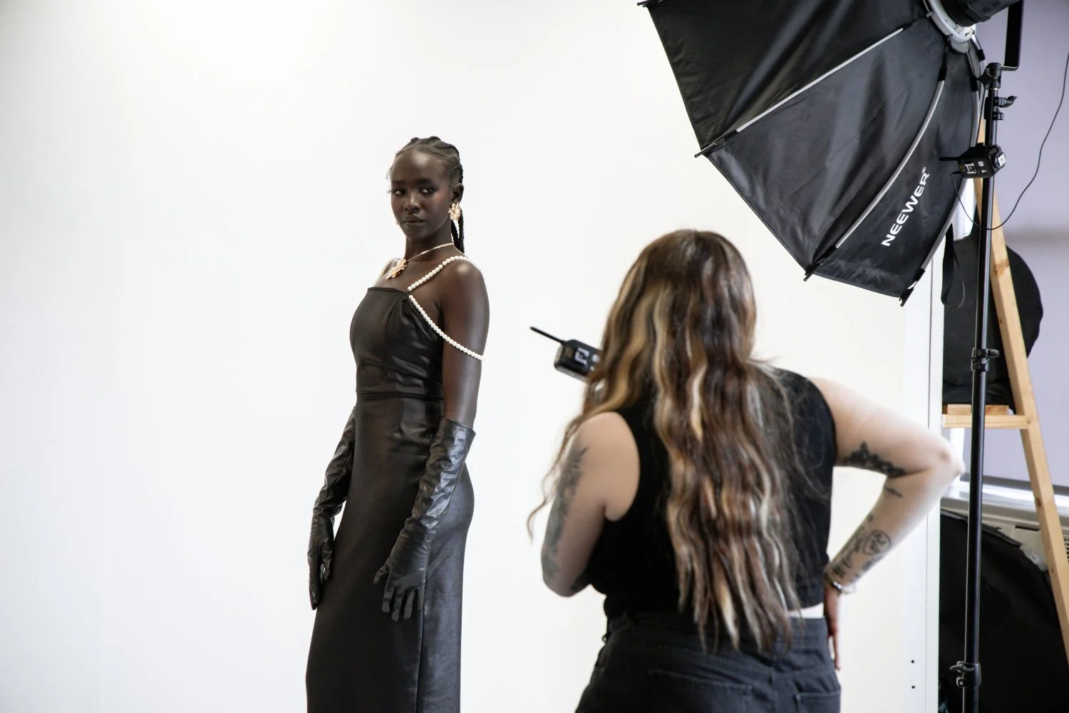 A woman in a black elegant dress with pearl jewelry and long black gloves posing in a photography studio, with a photographer and large studio lighting equipment visible.
