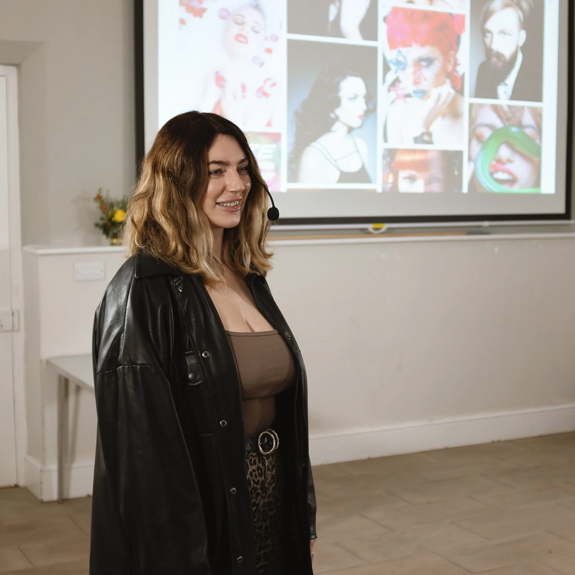 A woman with wavy brown hair and a lip piercing standing in front of a colorful collage of pictures, wearing a black leather jacket and patterned pants.