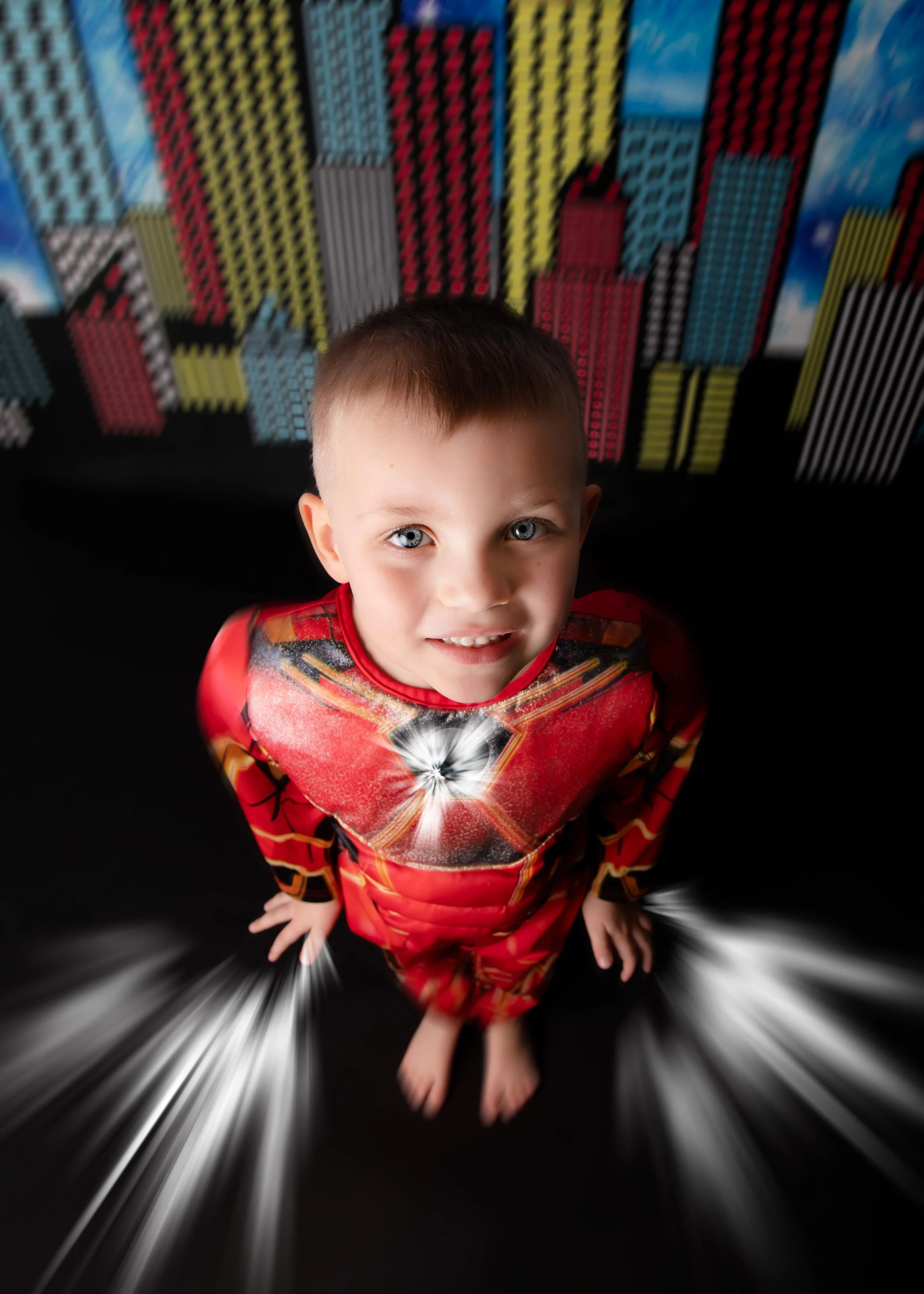 A young boy, dressed as a superhero in a red costume with a starburst design on his chest, stands barefoot on a black surface with a cityscape background, looking up at the camera with a bright expression.