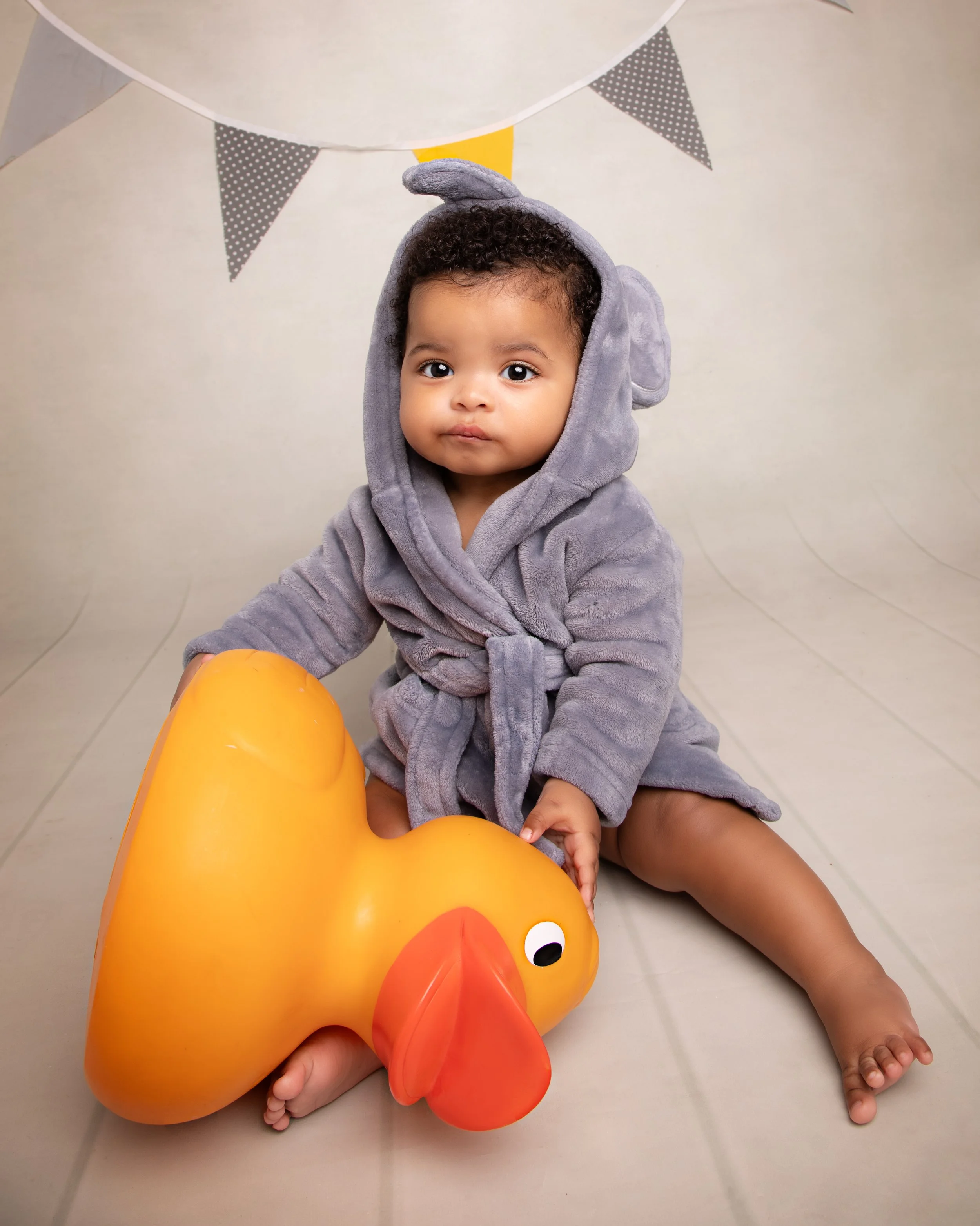 A young child wearing a gray elephant costume is sitting on the floor, holding a large orange rubber duck. The background has a neutral-colored wall with decorations including triangular flags hanging from a string.
