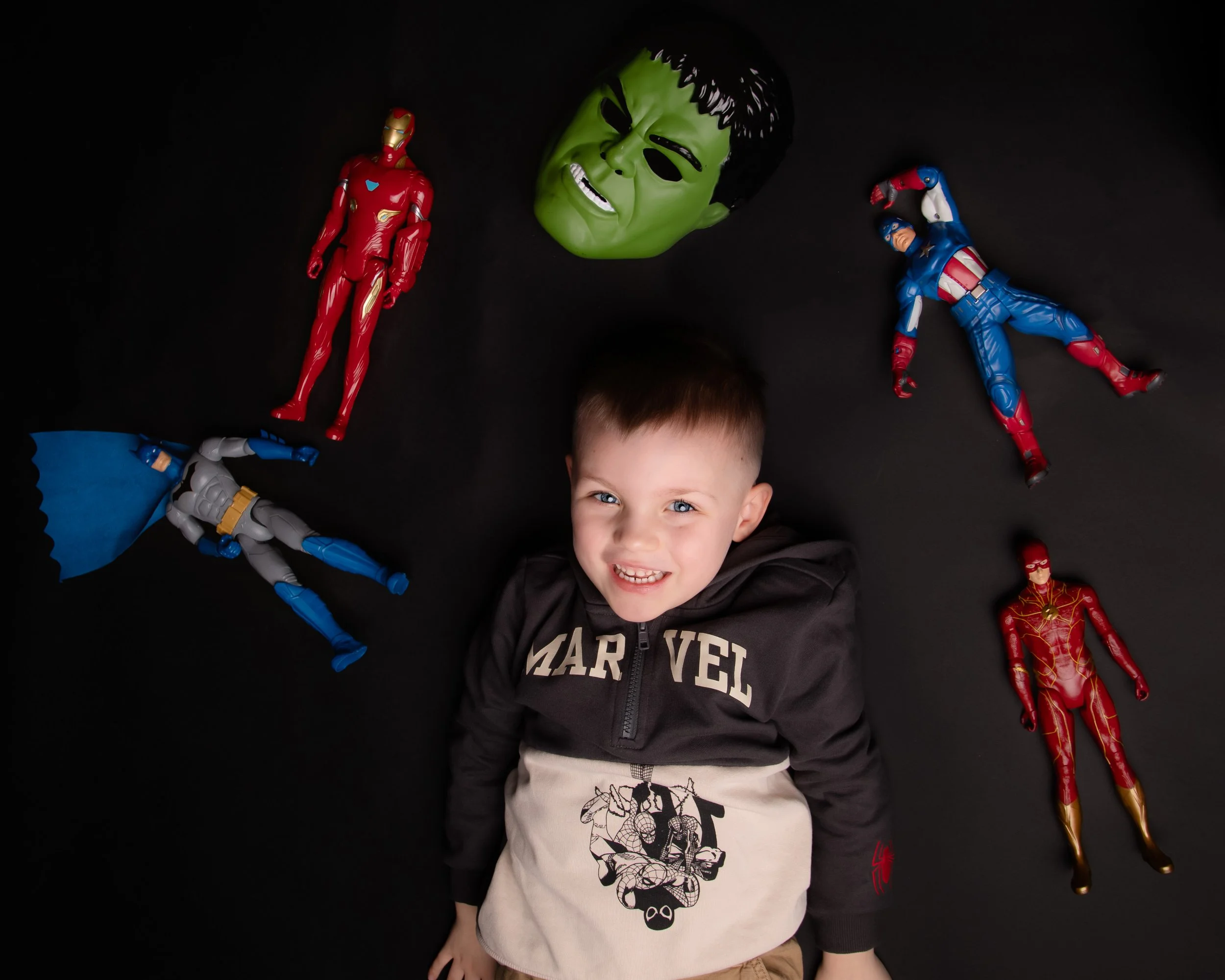 Young boy lying on black surface surrounded by Marvel superhero action figures and Hulk mask.