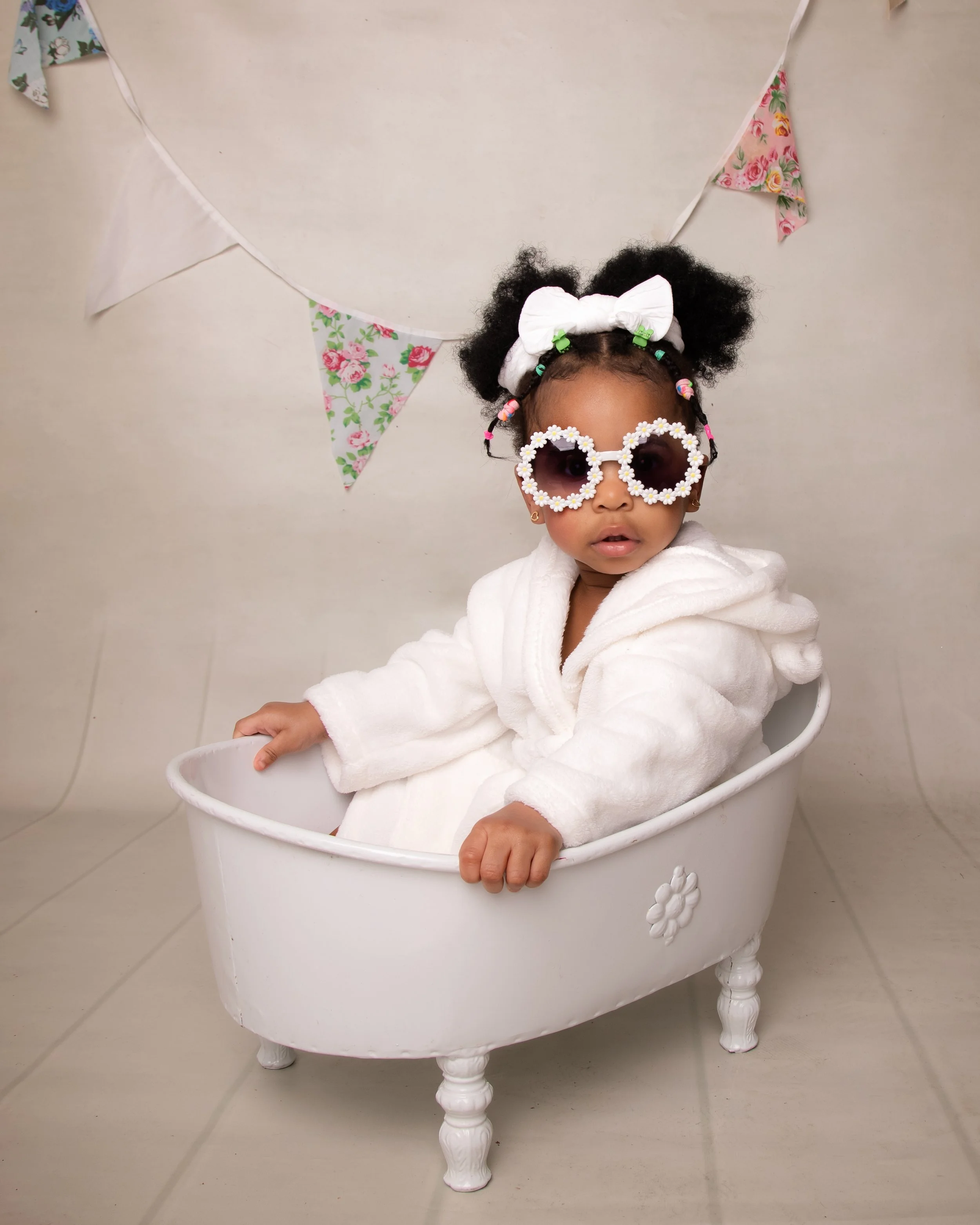 A young girl with black curly hair, wearing flower-shaped sunglasses, a white bow headband, and a white bathrobe, sitting in a small white bathtub with a floral decoration, in front of a beige wall with pink and floral bunting.