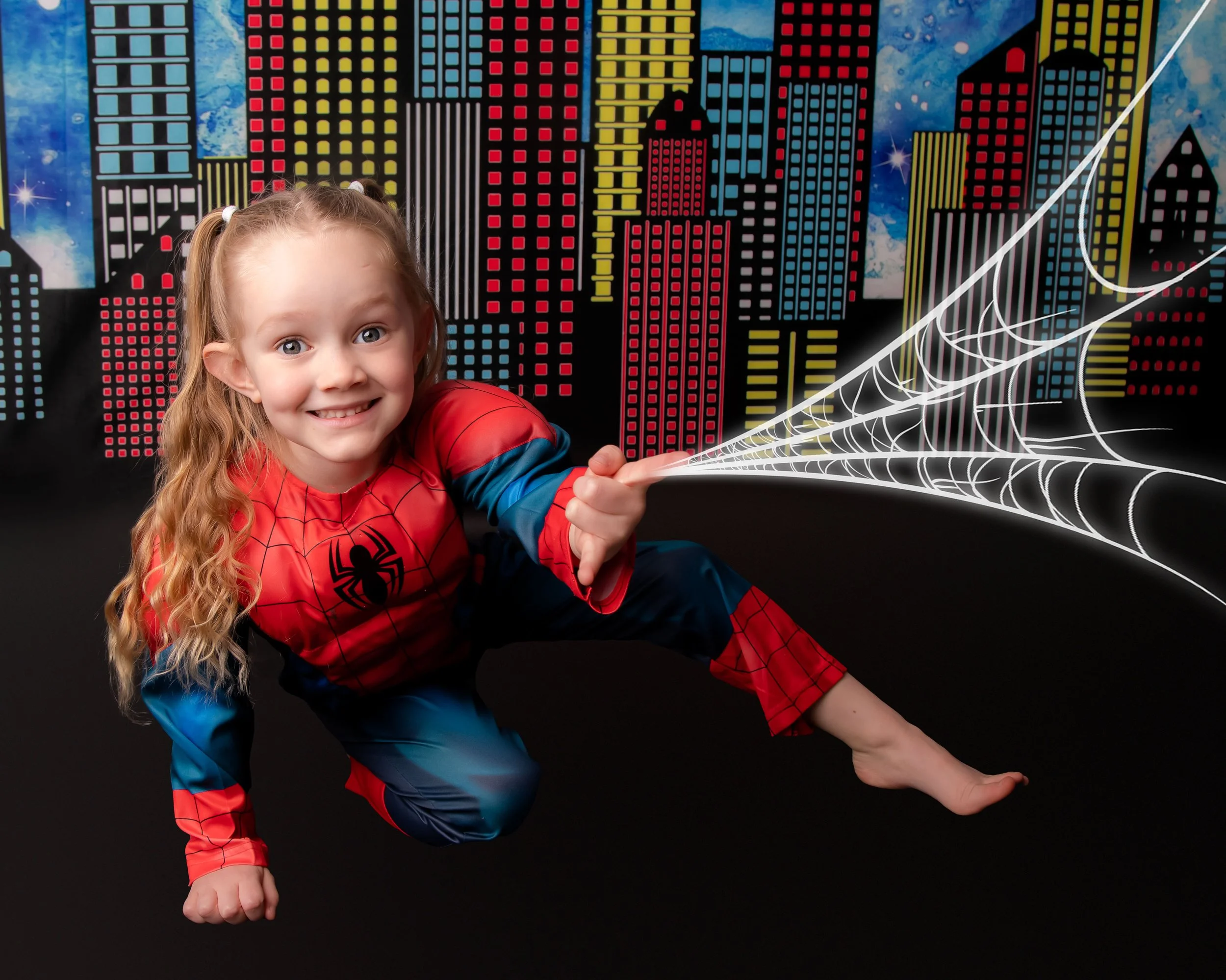 A young girl dressed as Spider-Man crouching in front of a colorful cityscape backdrop with glowing web details.