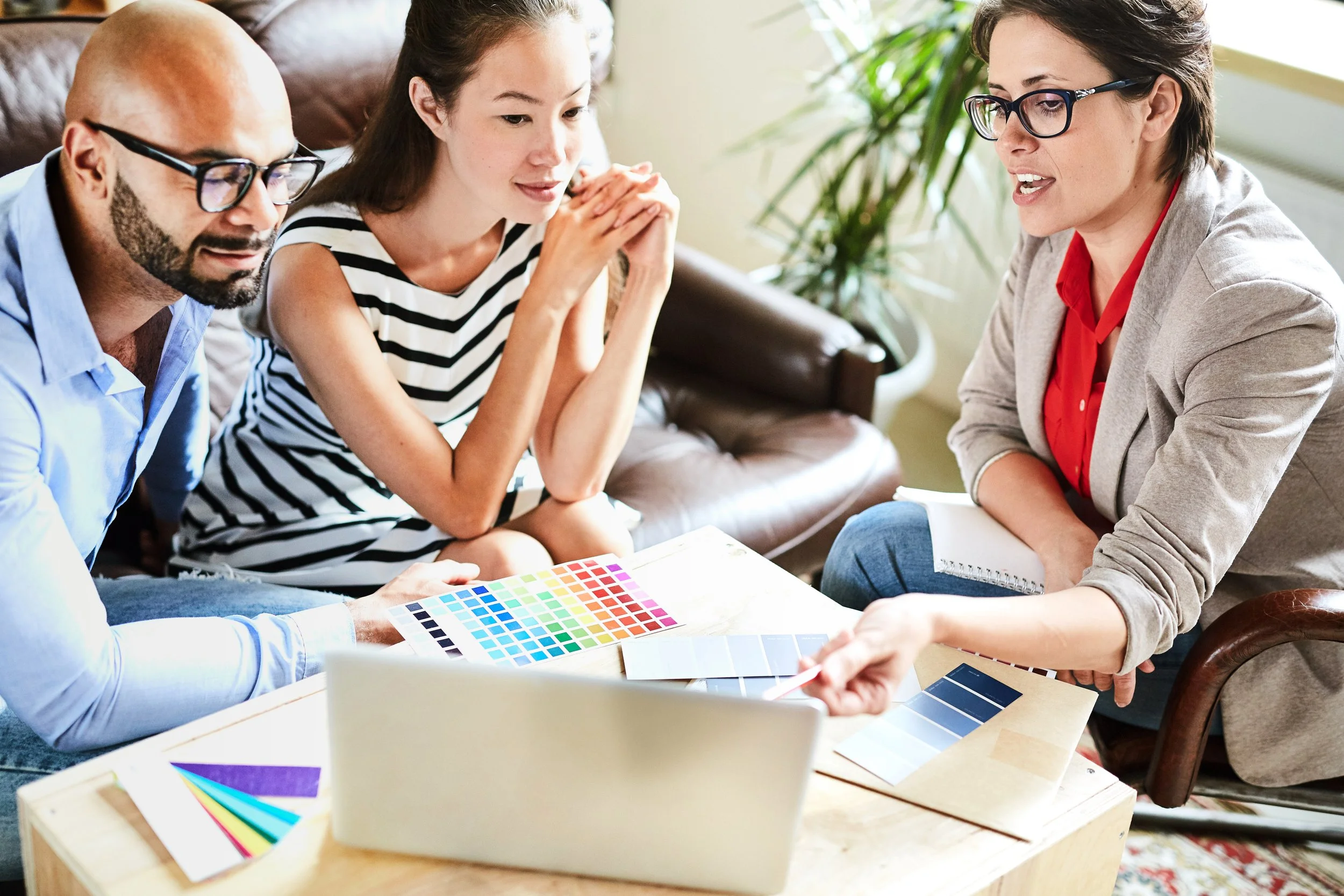 Three people discussing color swatches in a brightly lit room, with a laptop and color samples on the table.