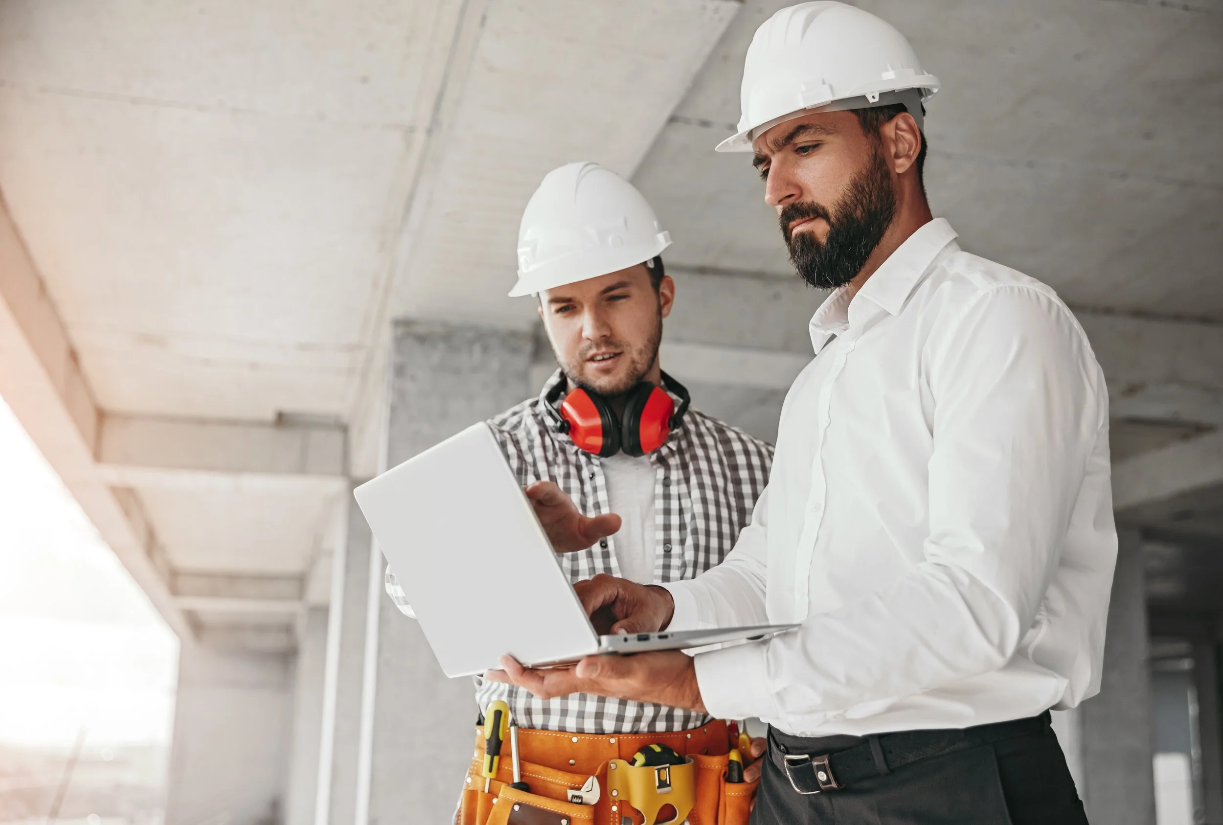 Two construction workers in hard hats reviewing plans on a laptop at a construction site.