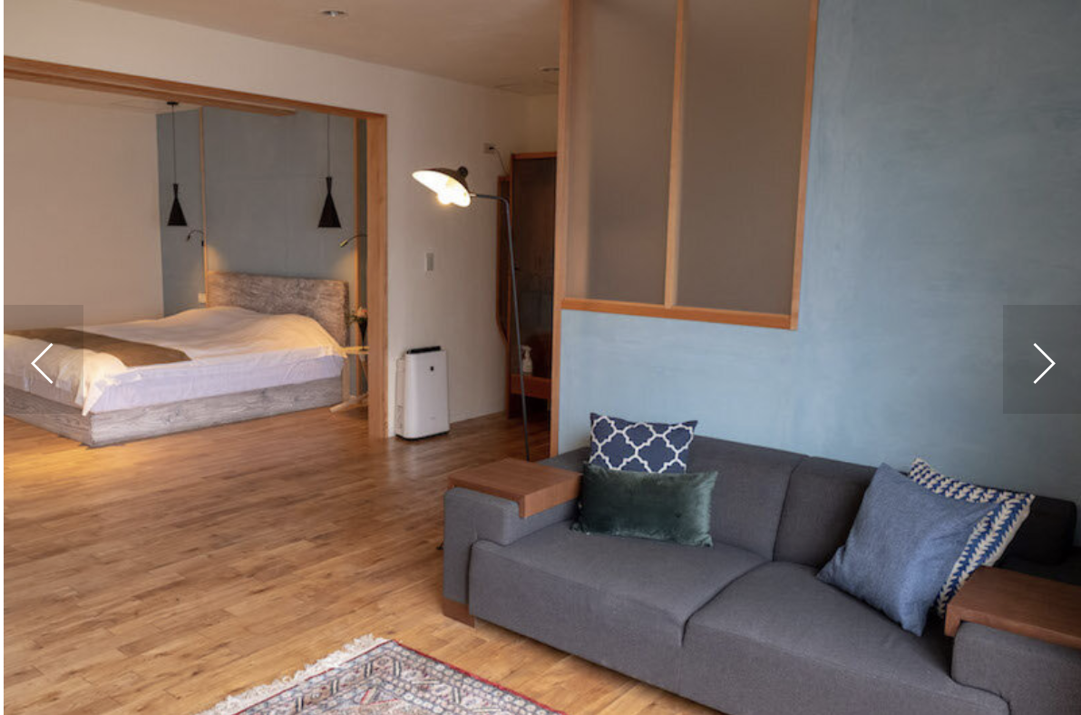 Modern living space with a gray sofa, decorative pillows, a bed in an alcove, hanging lamps, wooden floor, and a floor lamp.