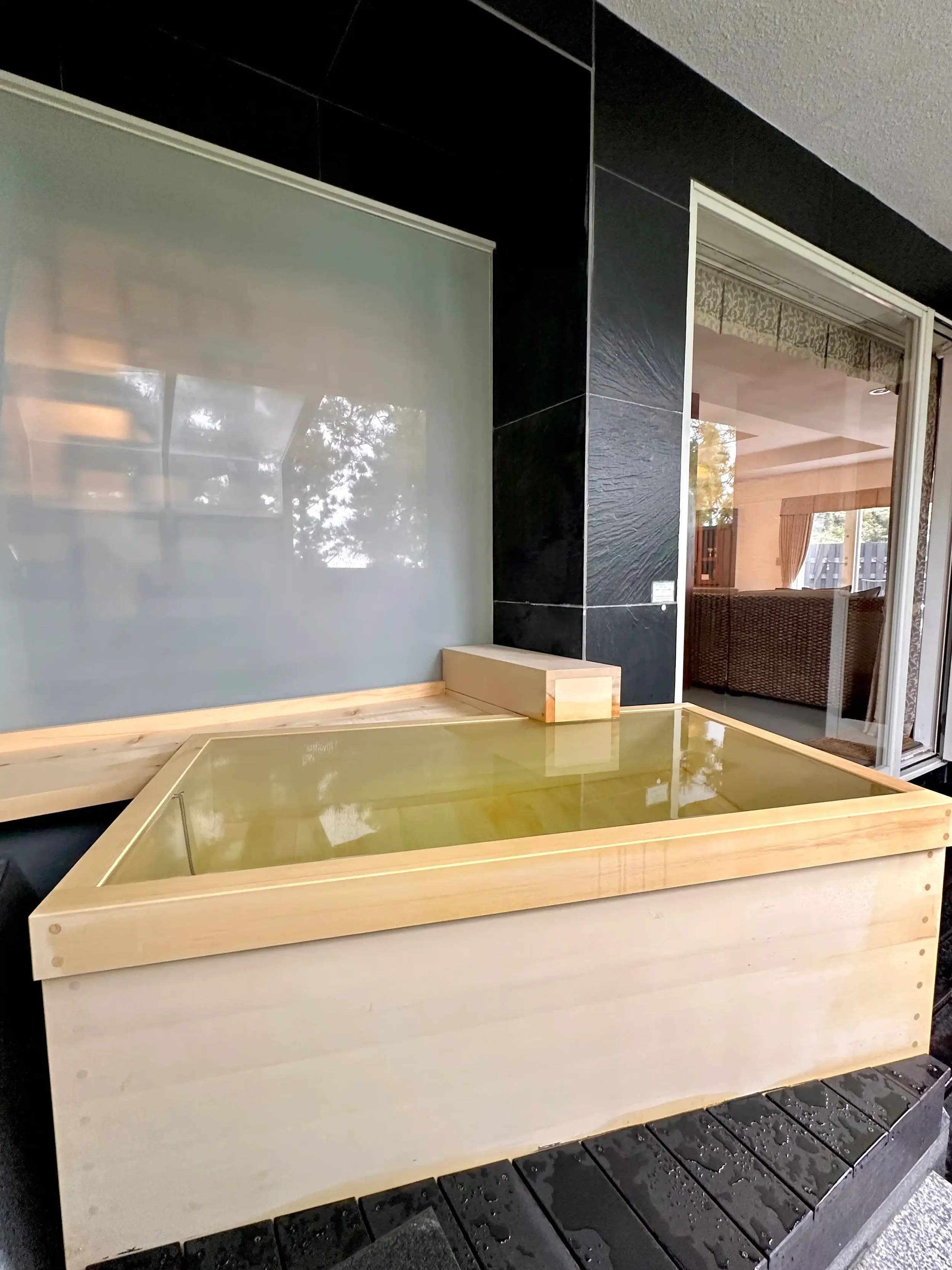 Interior view of a room featuring a black tiled bathtub and a glimpse of a bedroom with a bed, lamp, and curtains.