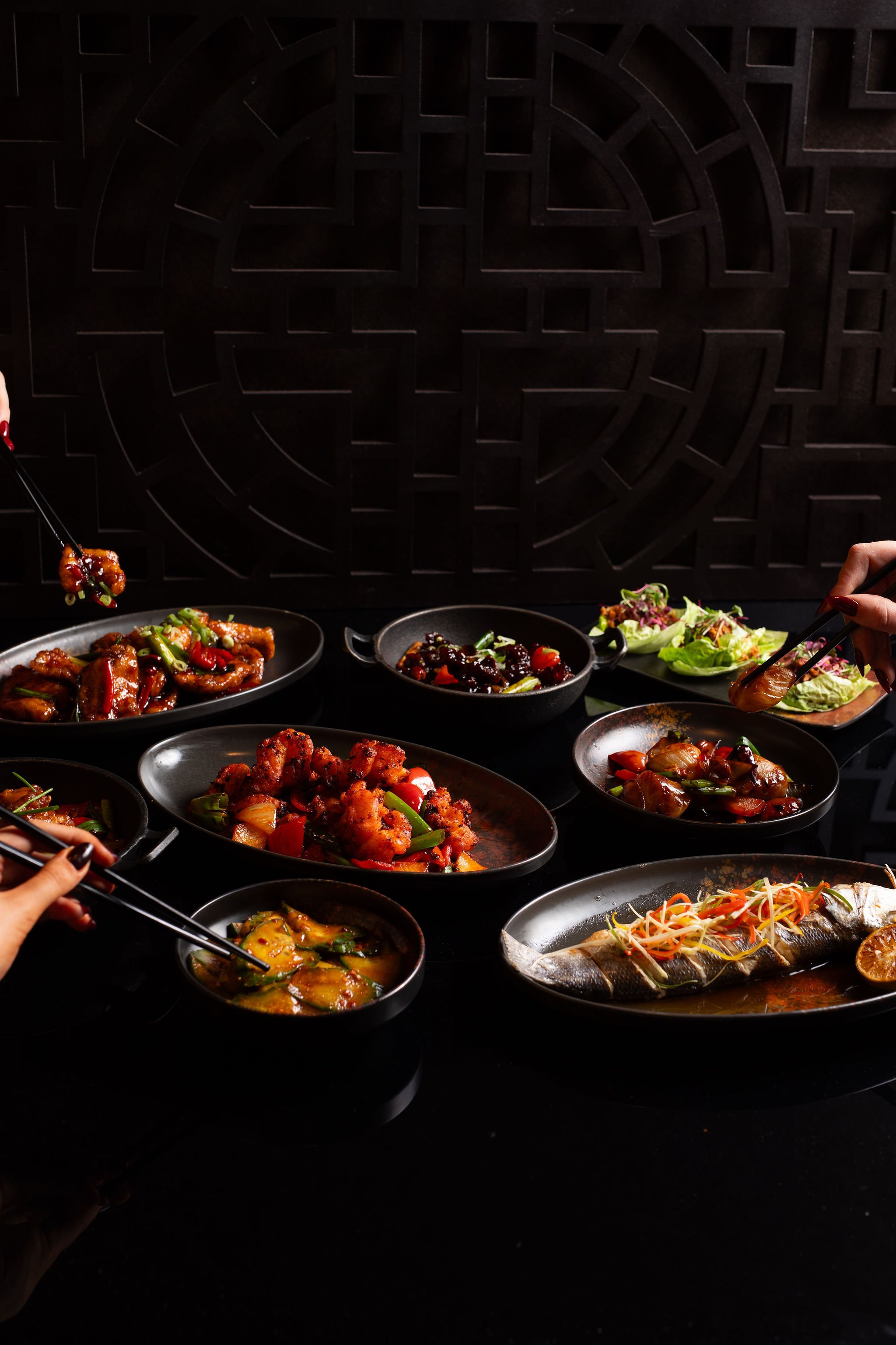 Several dishes of Asian cuisine, including a whole fish and various stir-fried dishes, on a black table with a dark background featuring a geometric pattern.