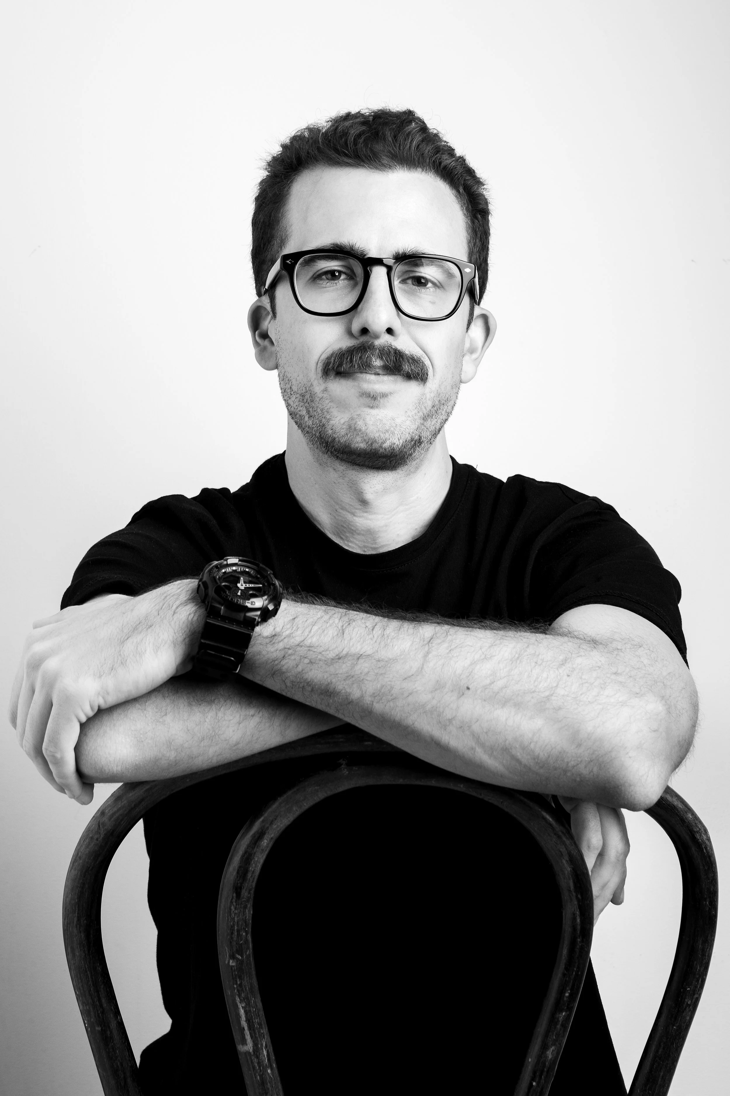 Hombre joven con barba, gafas grandes, camiseta negra, sentado con los brazos cruzados, en una silla de madera, fondo blanco.