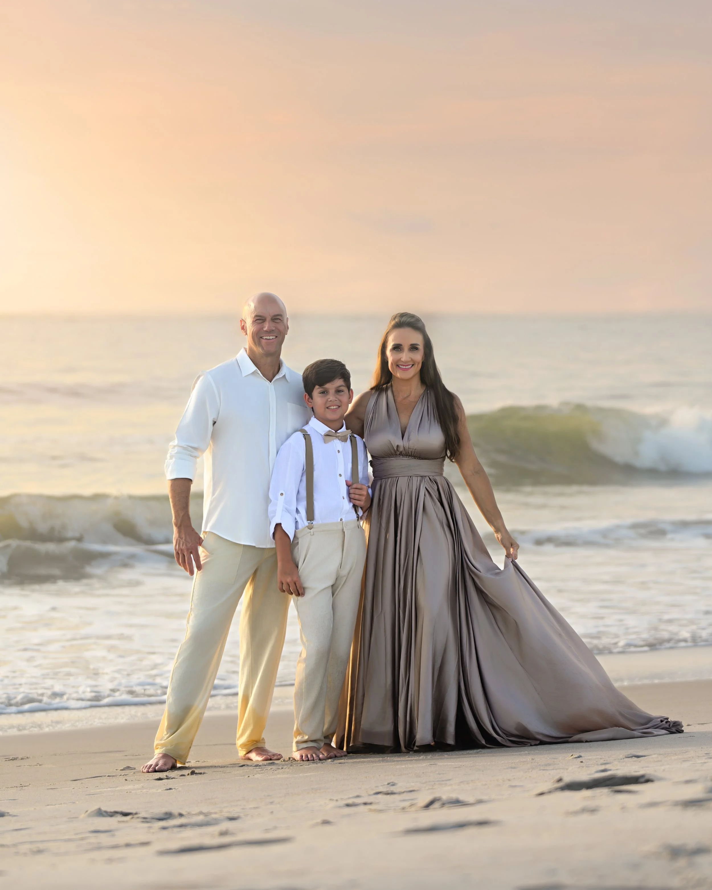 A family of four standing on a beach at sunset, smiling and dressed in formal attire. The father is wearing a white shirt and beige pants, the mother is in a long gray gown, the son is in a white shirt with suspenders, and the daughter is in a white shirt with a bow tie. They are standing close together with the ocean and a colorful sky in the background.