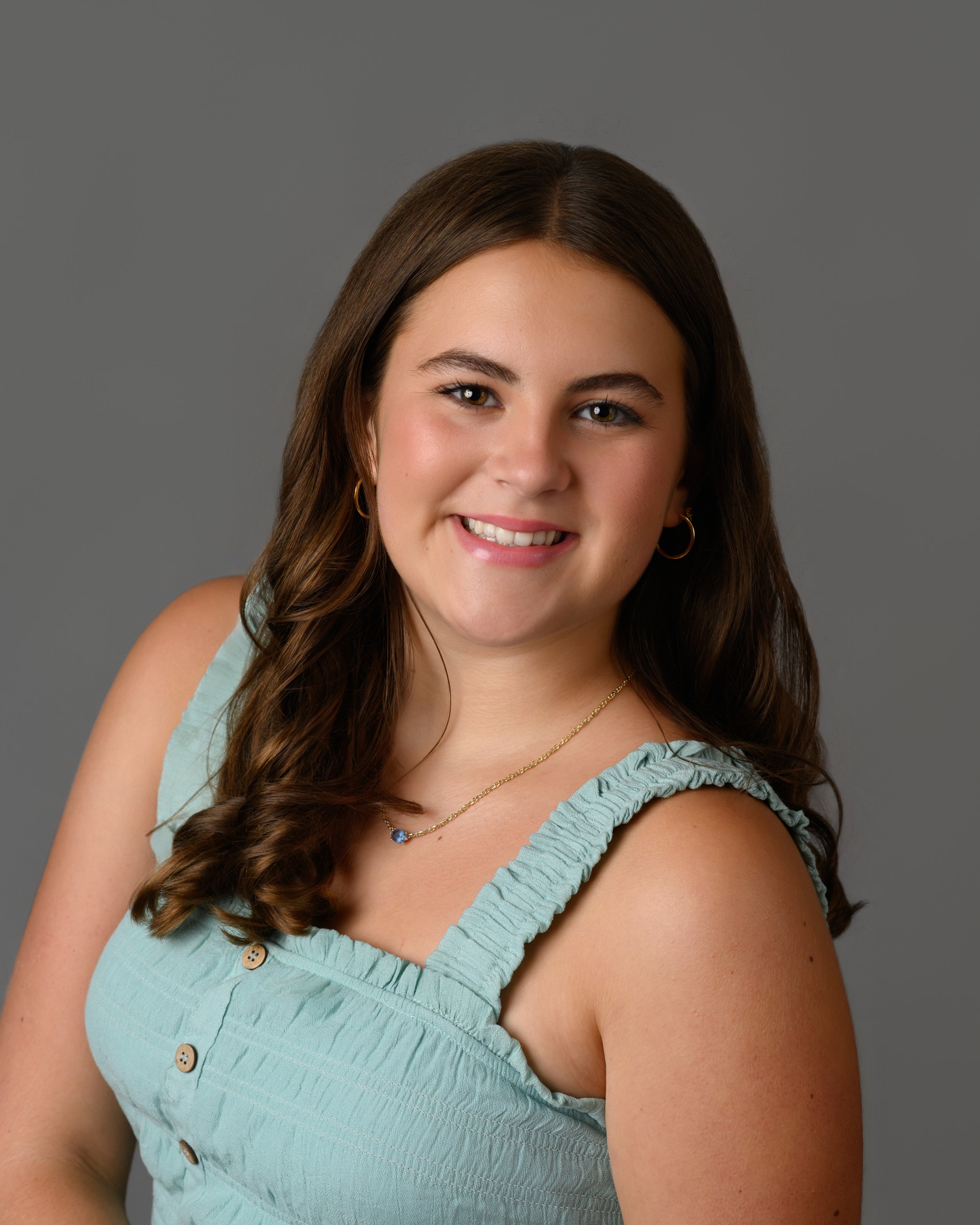 Close-up portrait of a young woman with long, wavy brown hair, smiling, wearing gold hoop earrings, a blue layered necklace with a blue stone pendant, and a sleeveless light blue top, against a gray background.