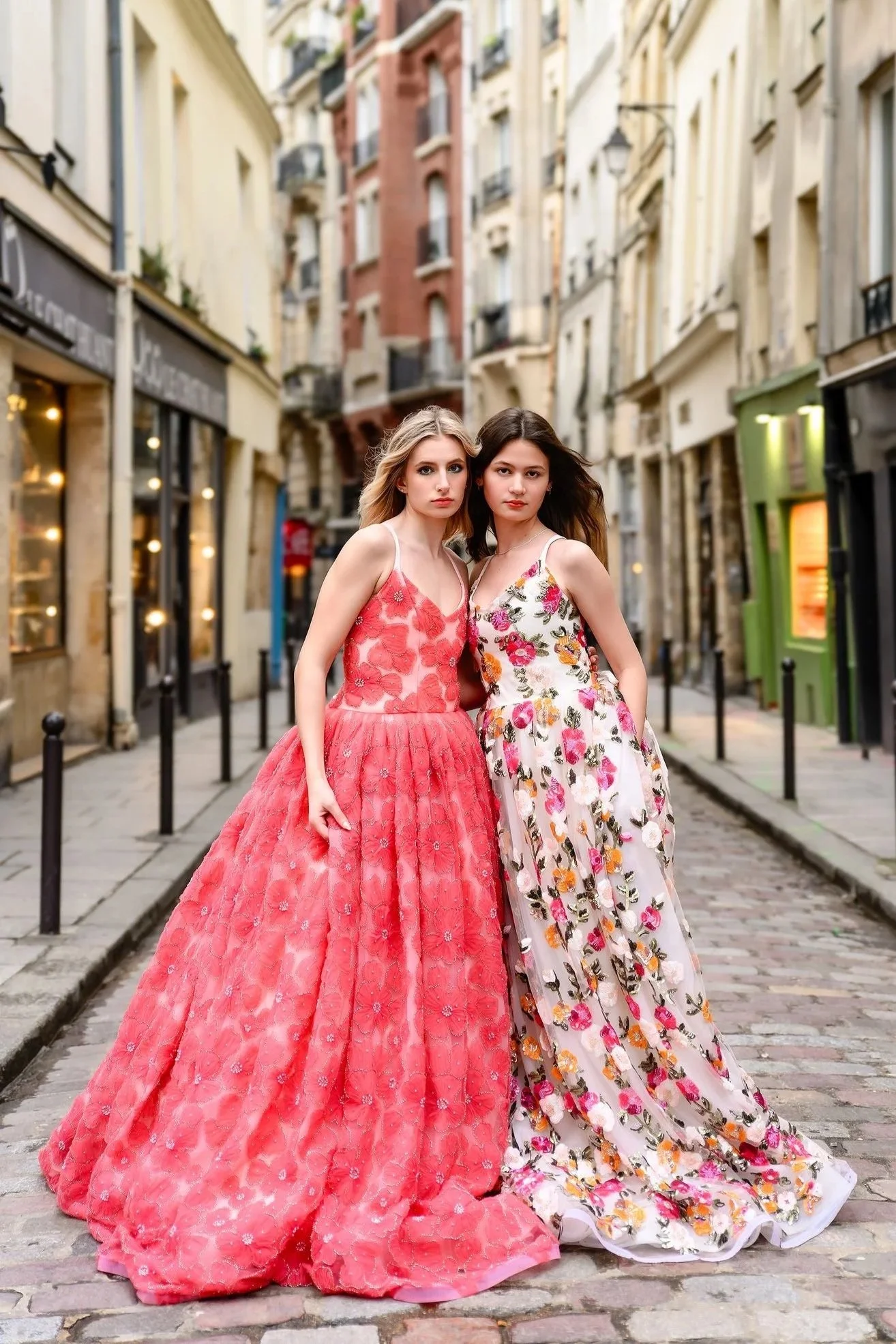 Two young women in elaborate floral dresses standing close together on a cobblestone street in an urban area, with buildings lining both sides in the background.