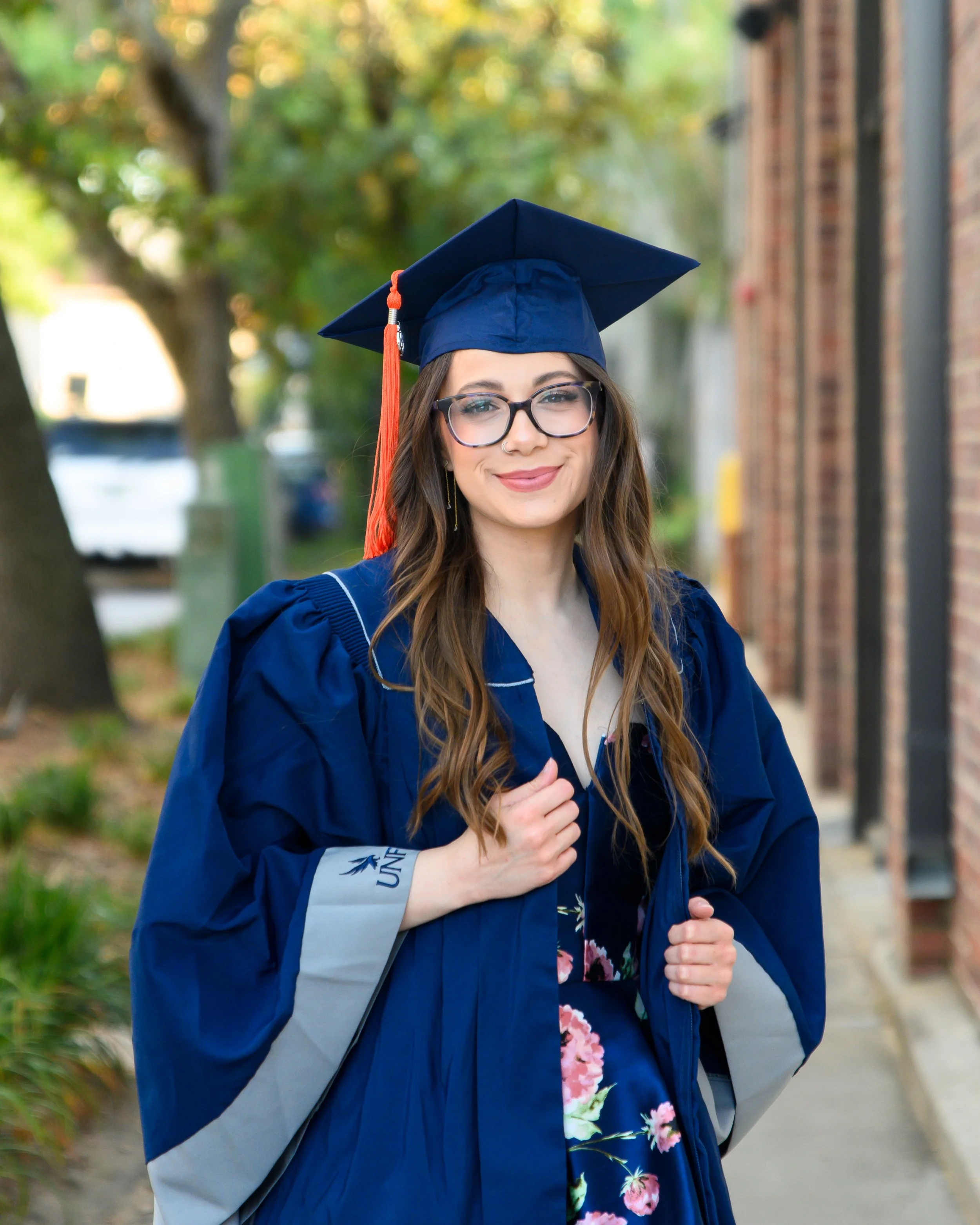 A young woman wearing glasses and a blue graduation cap and gown, smiling outdoors with trees and a brick building in the background.