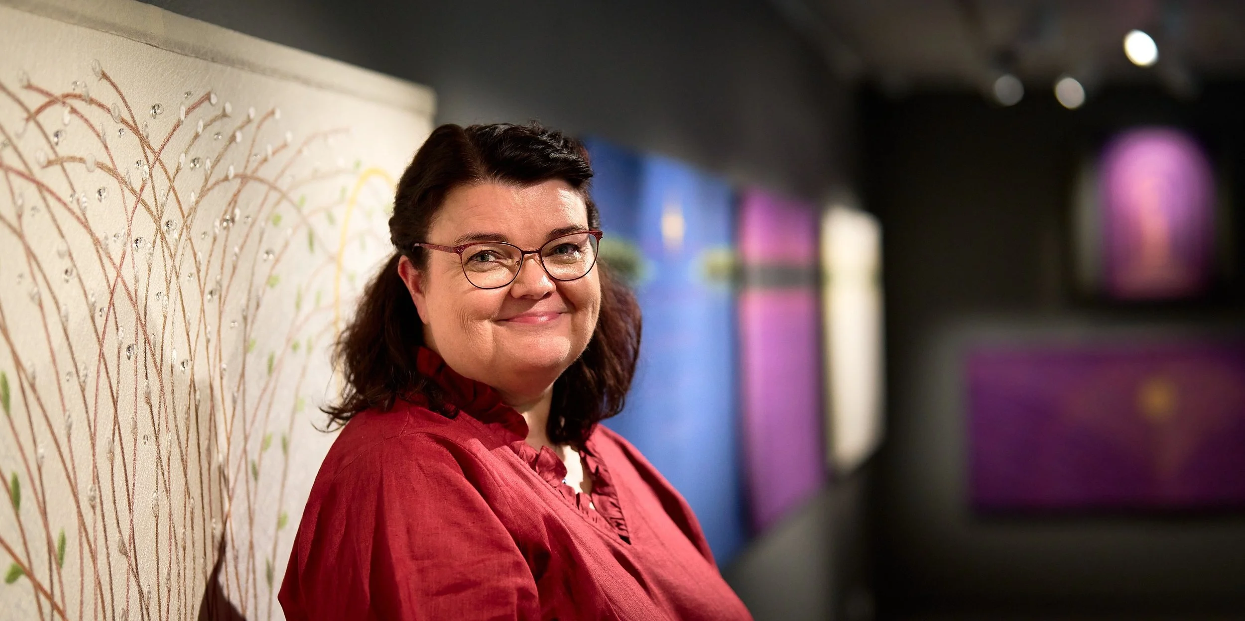 Helena seisoo kirkkotekstiilitöidensä esillä näyttelyssään.