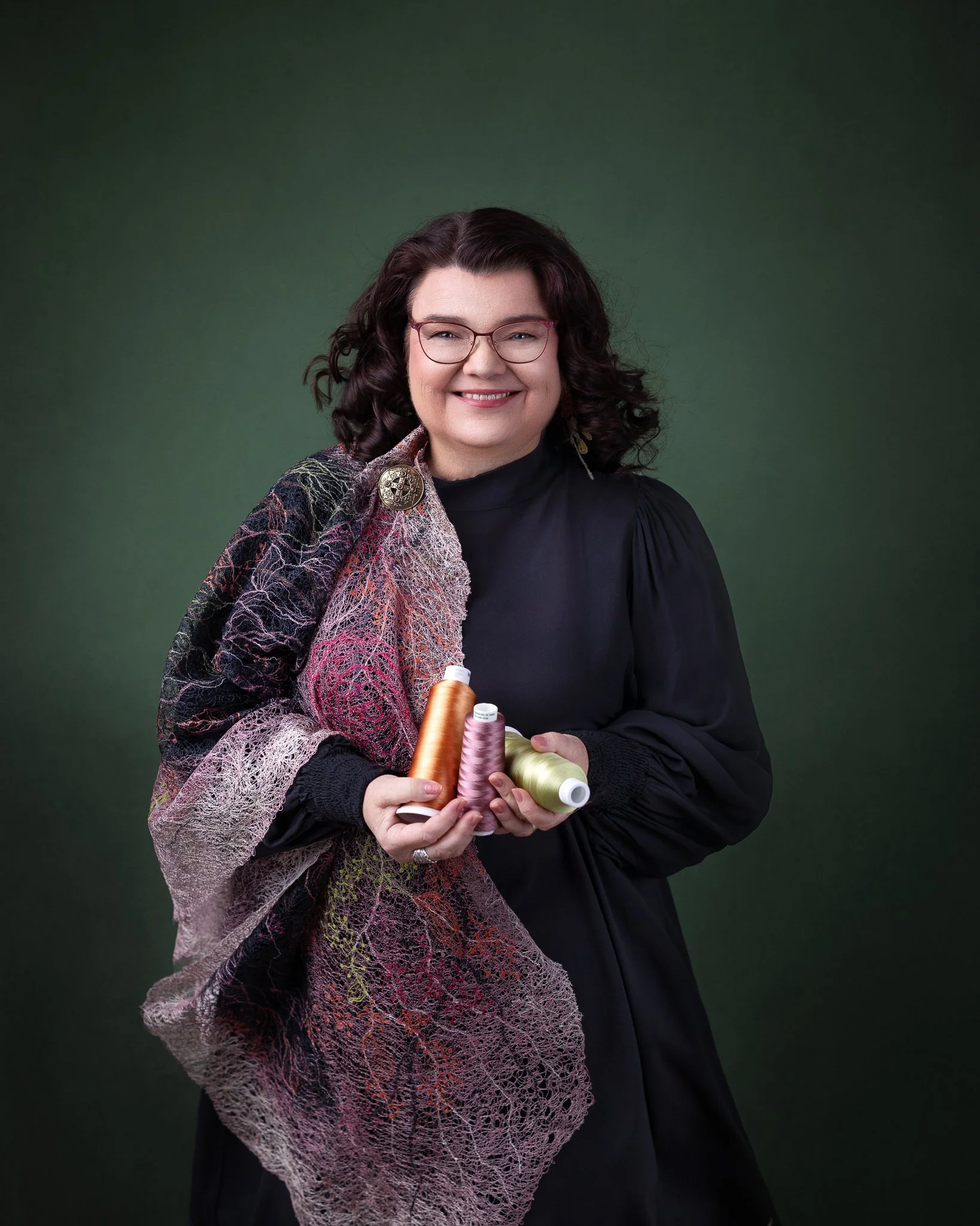 Helena seisoo suoraan kameraa kohti, pitelee käsissään lankakeriä ja olkapäillä hänen ommeltu työnsä, taustalla tummanvihreä seinä.