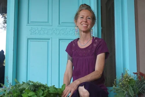 Woman smiling and sitting on steps in front of a bright blue door, with plants around her.