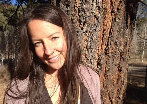 A smiling woman with long brown hair standing outdoors near a tree.