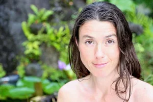 A woman with wet hair and no shirt in an outdoor garden setting with green foliage and purple flowers.