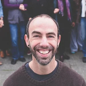 Close-up of a smiling man with a beard and short hair, wearing a dark sweater, with people in the background.