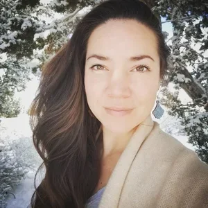A woman with long dark hair taking a selfie outdoors with trees in the background.