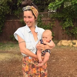 A woman holding a baby outdoors with a wooden fence and greenery in the background.
